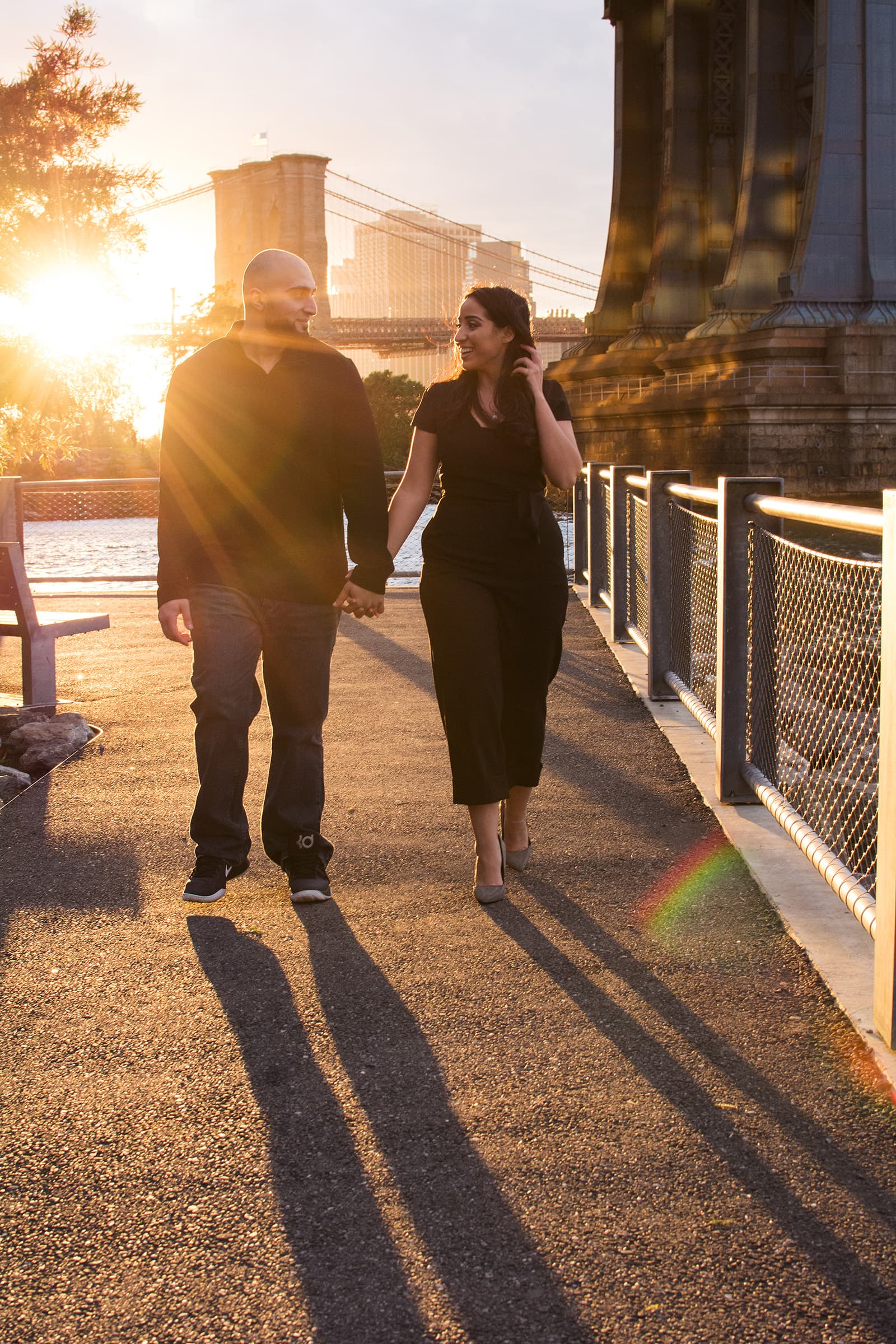 Couple walking hand in hand and smiling during a relaxed golden-hour portrait session beside the waterfront and bridge.