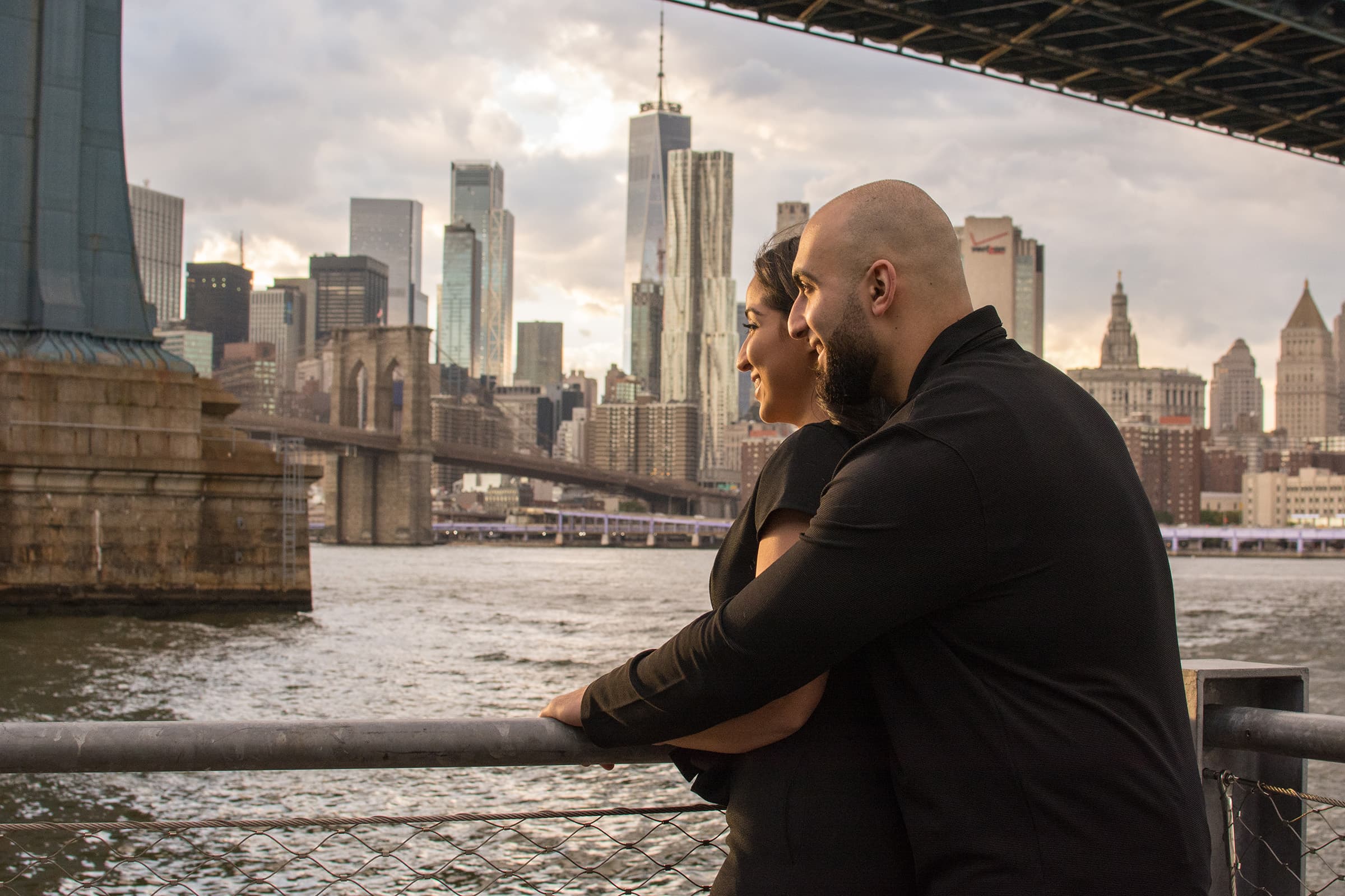 Couple embracing beside the waterfront while looking toward the city skyline during a relaxed urban portrait session.