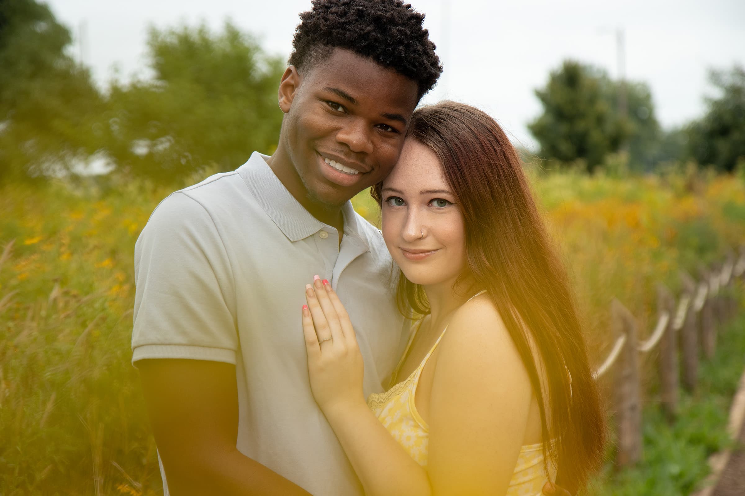 Smiling couple posing together in a garden setting during a natural outdoor portrait session with soft yellow flowers.