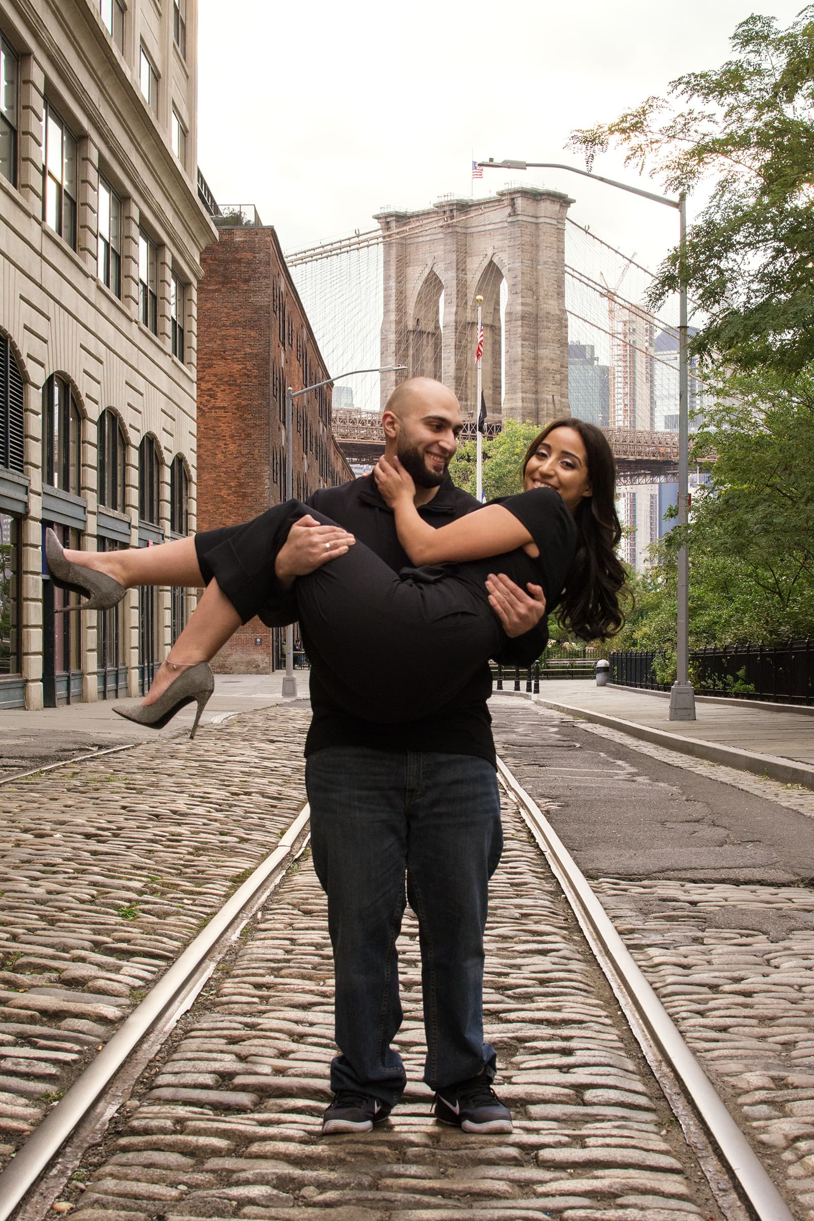 Playful couple smiling as the man carries his partner along a cobblestone street with a suspension bridge behind them.
