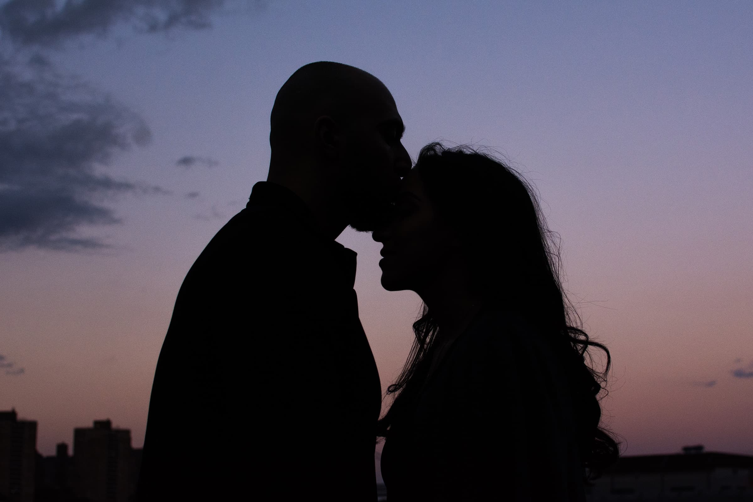 Romantic couple touching foreheads in silhouette against a soft purple sunset sky during an intimate portrait session.