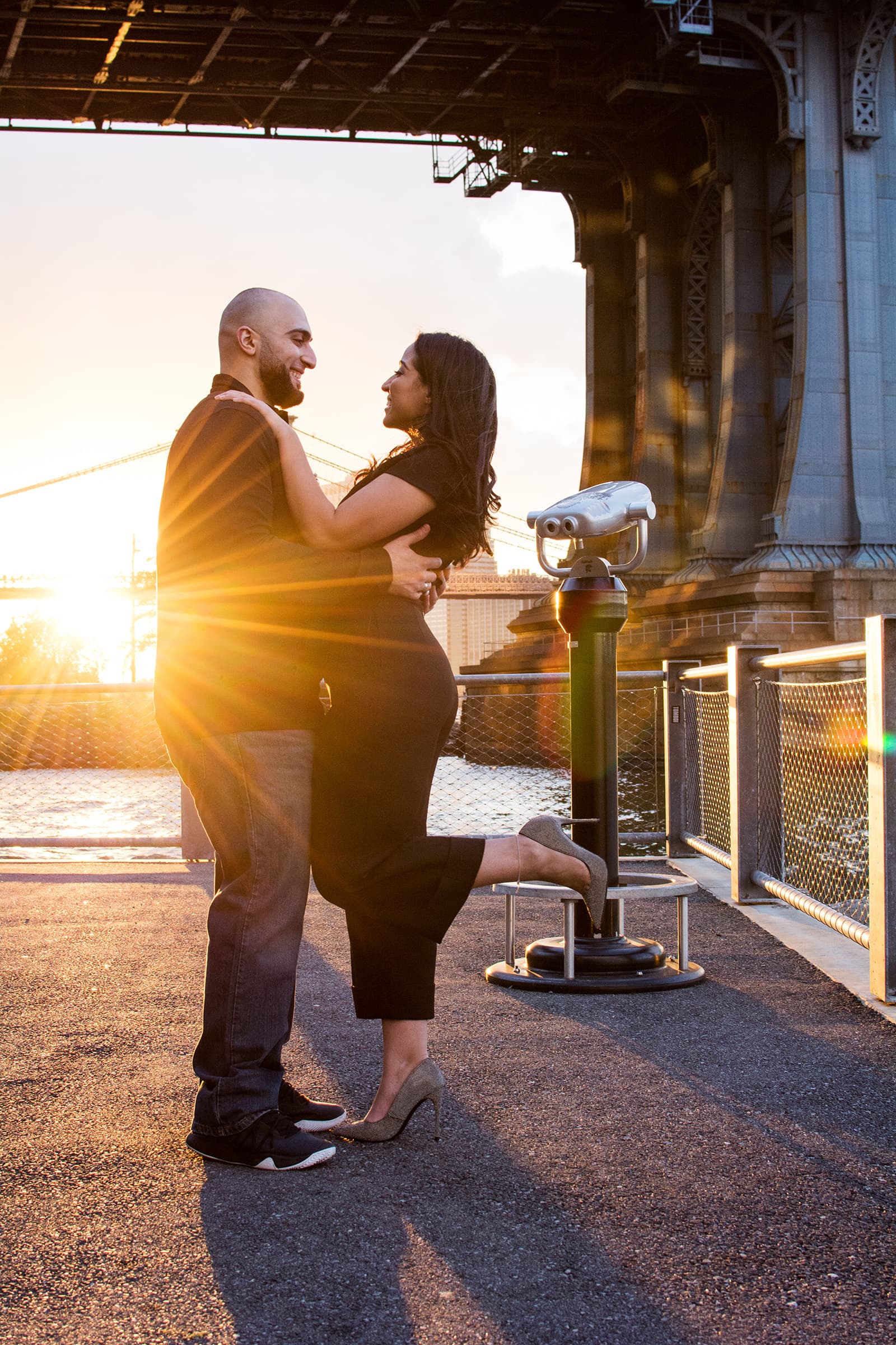 Couple embracing and smiling together beside the waterfront as warm golden sunlight creates a dramatic portrait backdrop.
