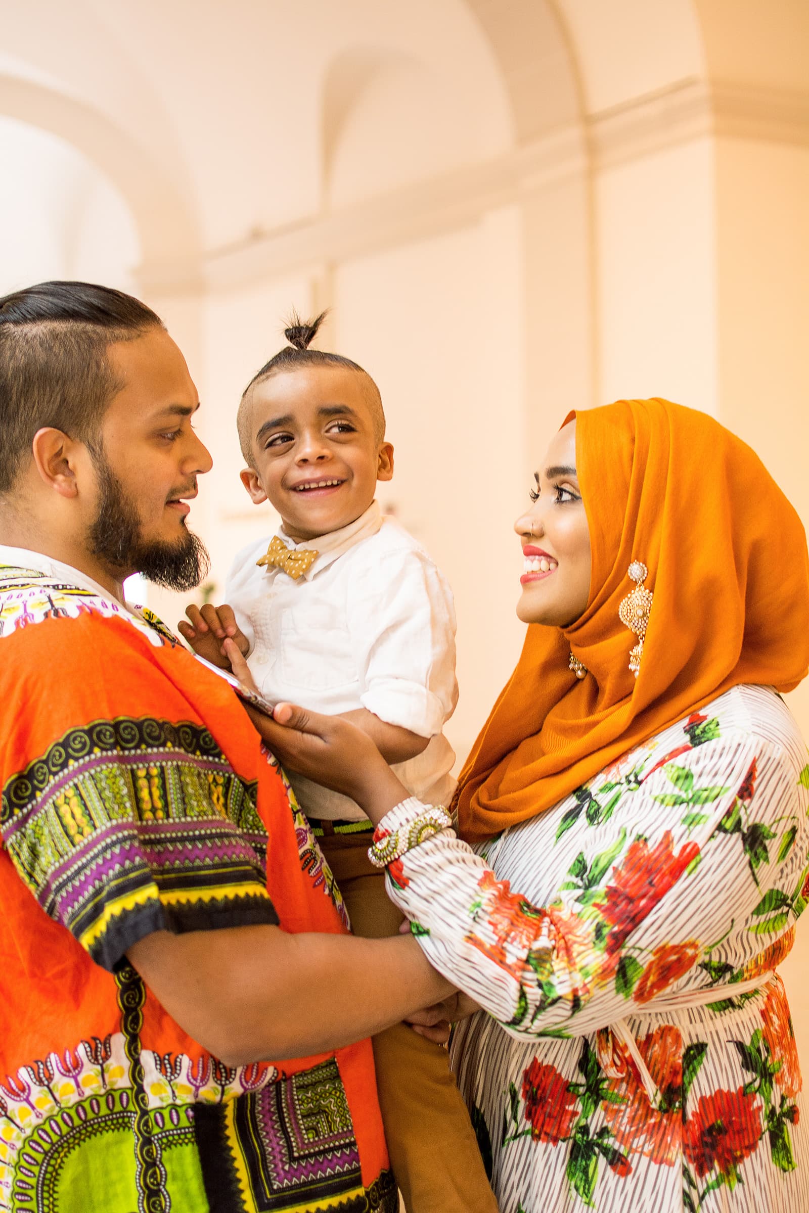 Muslim family portrait of a mother in an orange hijab, father, and smiling young son photographed together indoors.