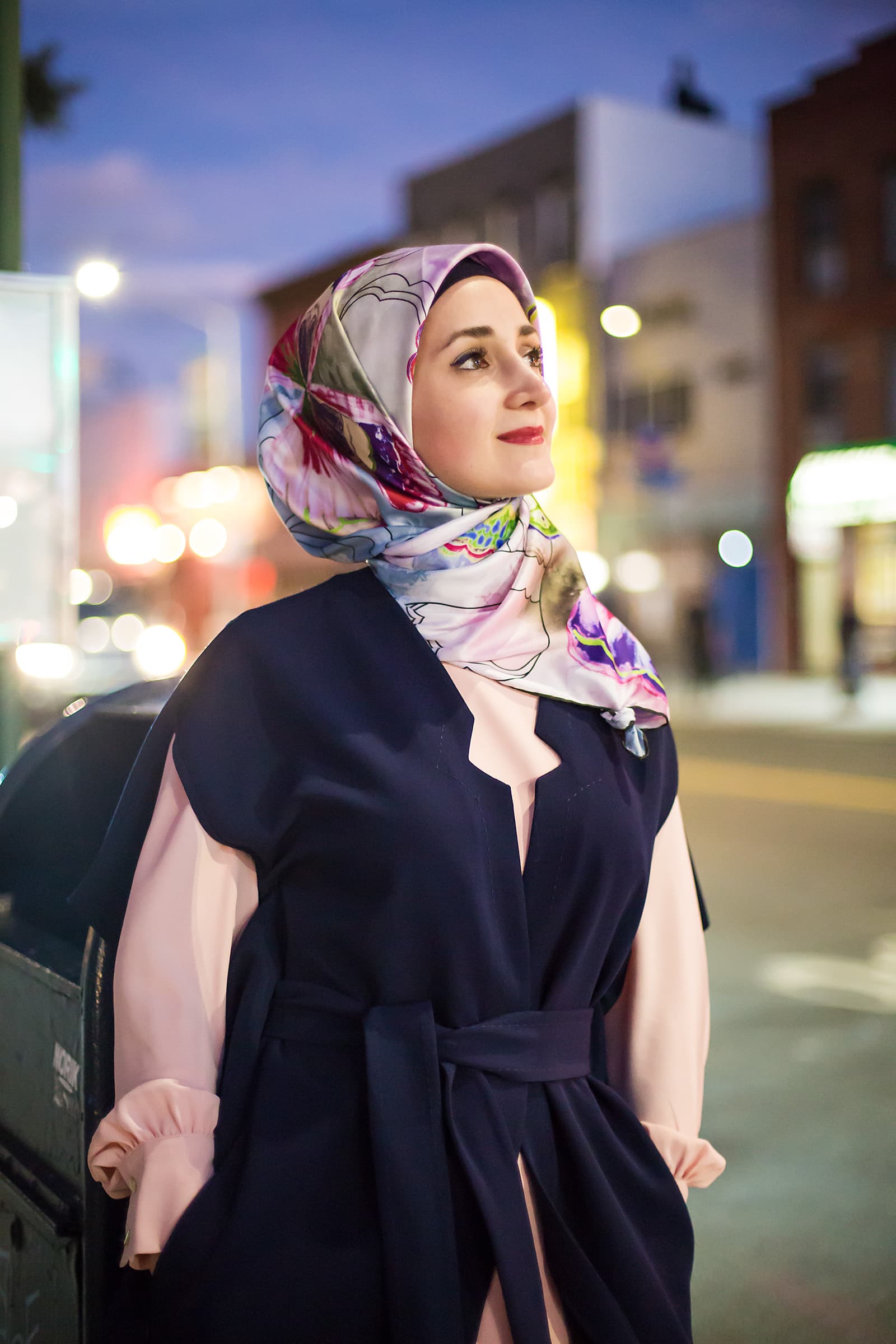 Evening street portrait of a Muslim woman in a colorful floral hijab and navy outfit with soft city lights behind her.