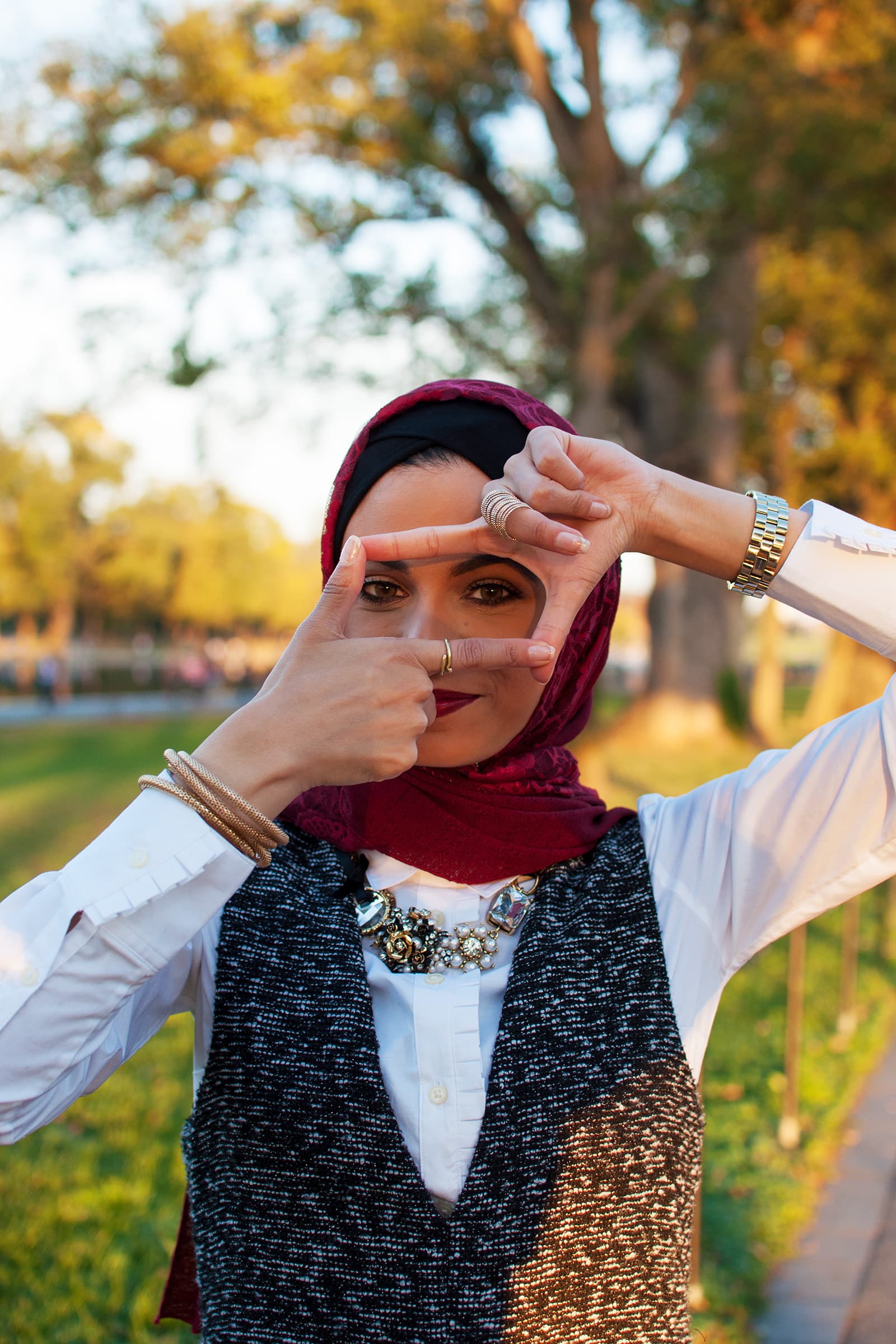 Creative portrait of a Muslim woman in a burgundy hijab framing her eyes with her hands in a sunlit public park.