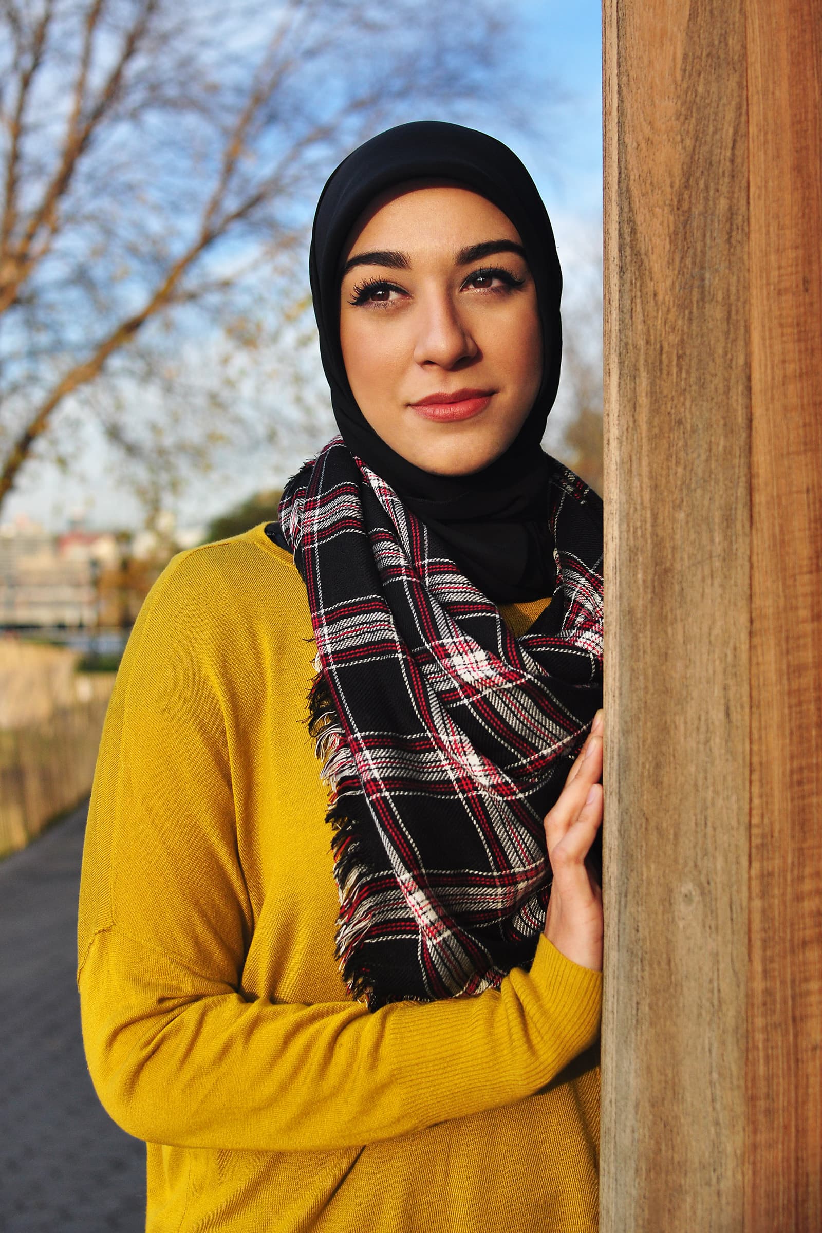 Muslims of America portrait of a Muslim woman in a black hijab and mustard sweater standing outdoors beside a wooden post.