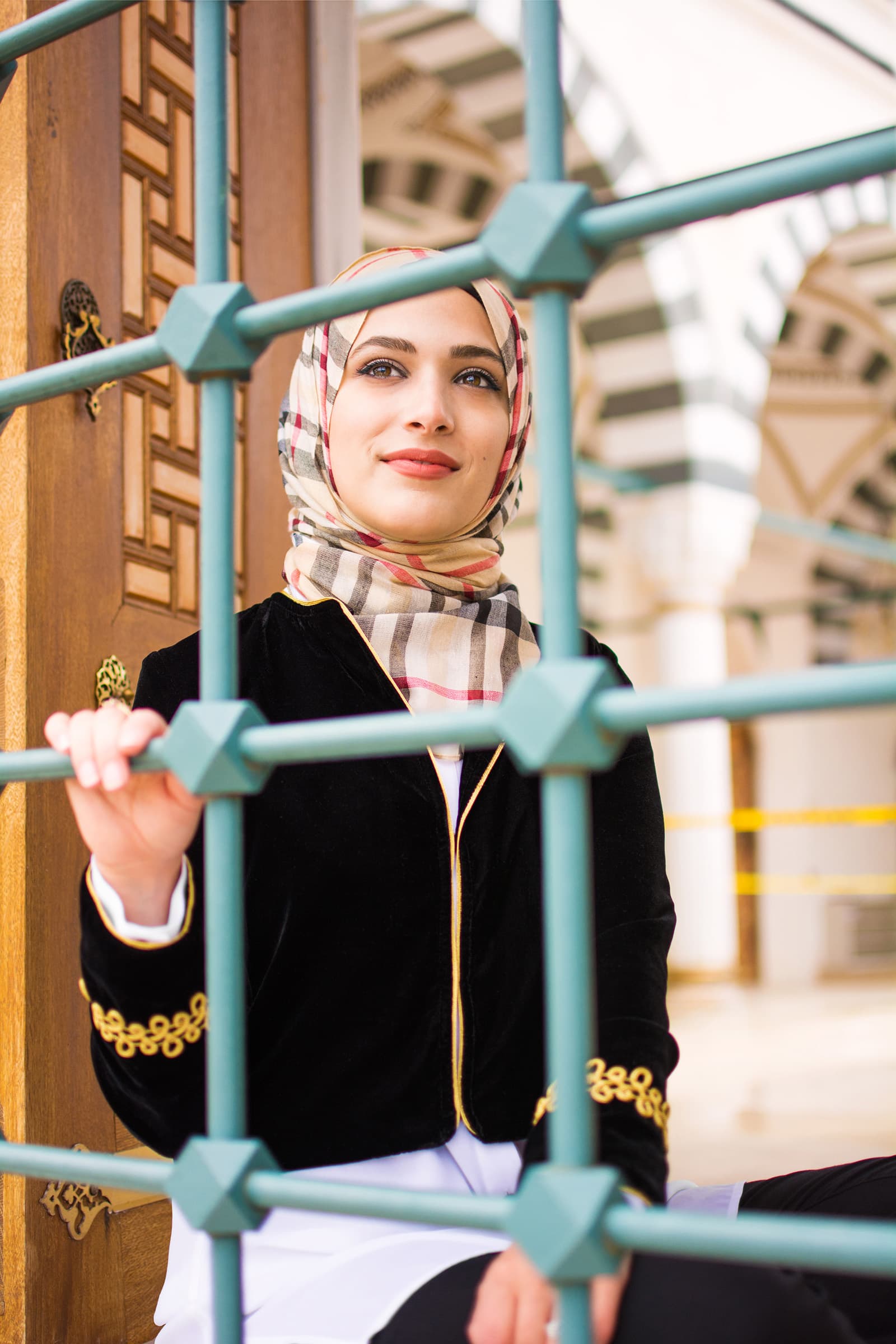 Portrait of a Muslim woman in a patterned hijab and black jacket framed by architectural metalwork outside a mosque.