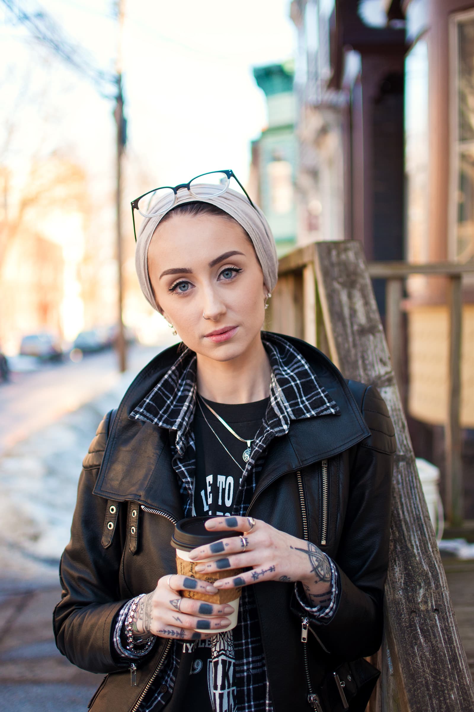 Young Muslim woman in a neutral head wrap and black leather jacket holding coffee during an outdoor urban portrait session.