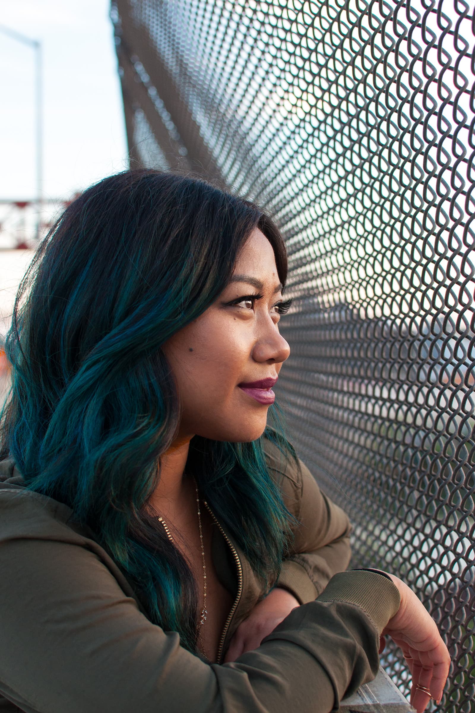 Profile portrait of a Muslim woman with teal hair leaning beside a chain-link fence during an outdoor urban photo session.