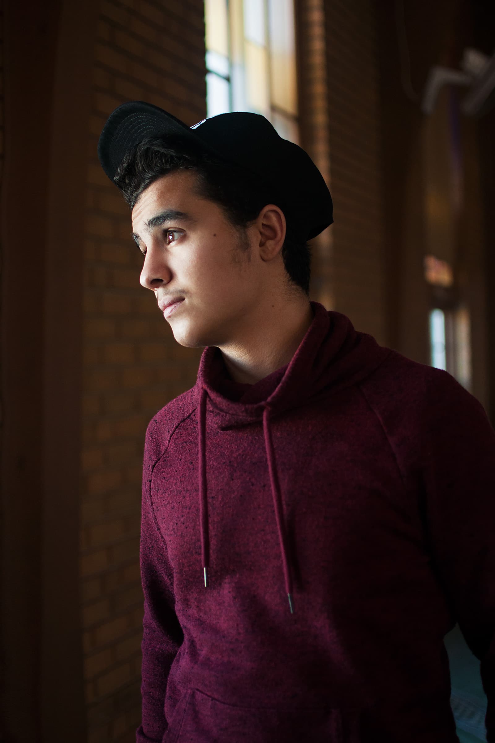 Muslims of America indoor portrait of a young Muslim man in a burgundy hoodie and black cap beside soft window light.