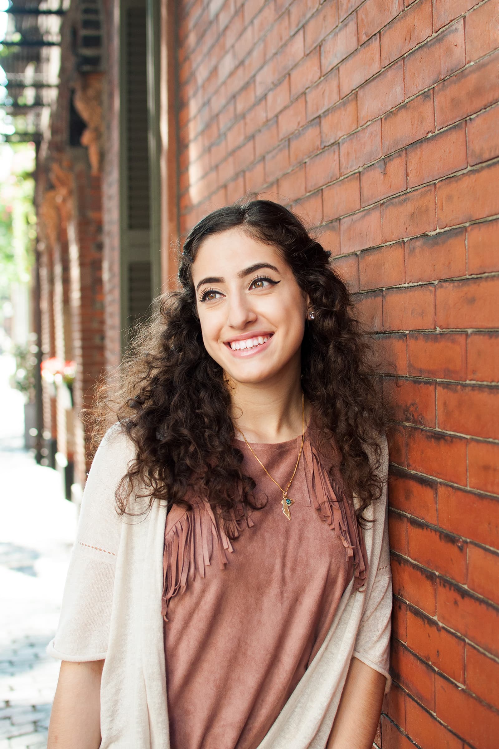 Muslims of America portrait of a smiling young Muslim woman with long curly hair standing beside a historic brick wall.