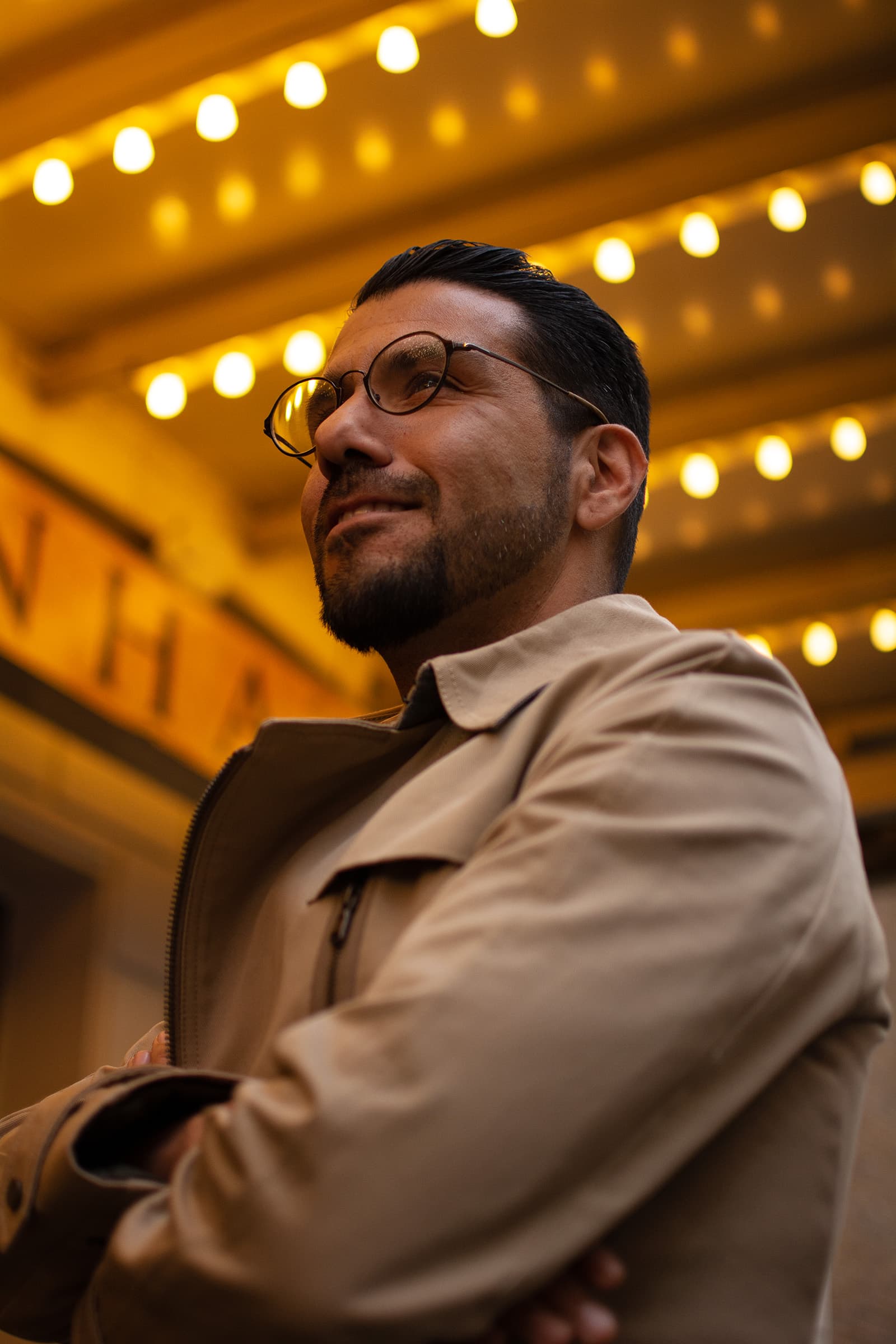 Muslims of America portrait of a Muslim man with glasses and beard beneath warm marquee lights in an urban setting.