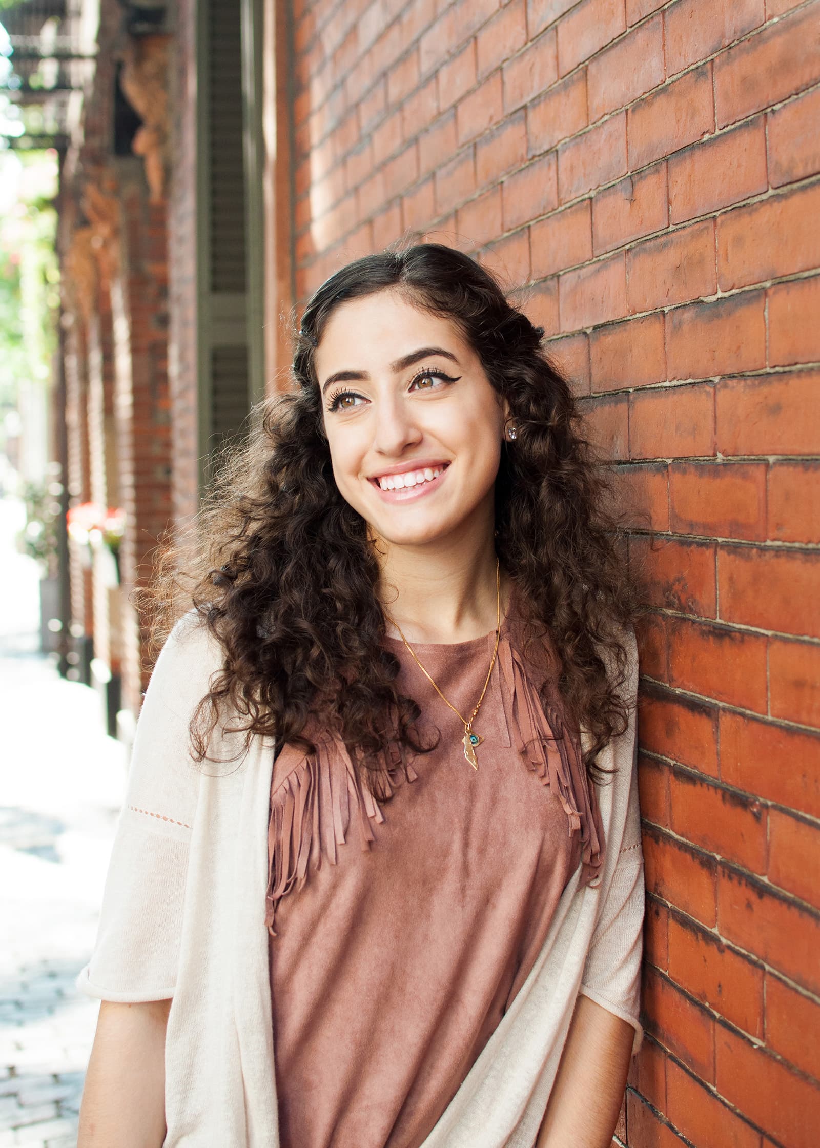 Natural-light portrait of a smiling woman with long curly hair posing beside a red brick wall in an urban setting.
