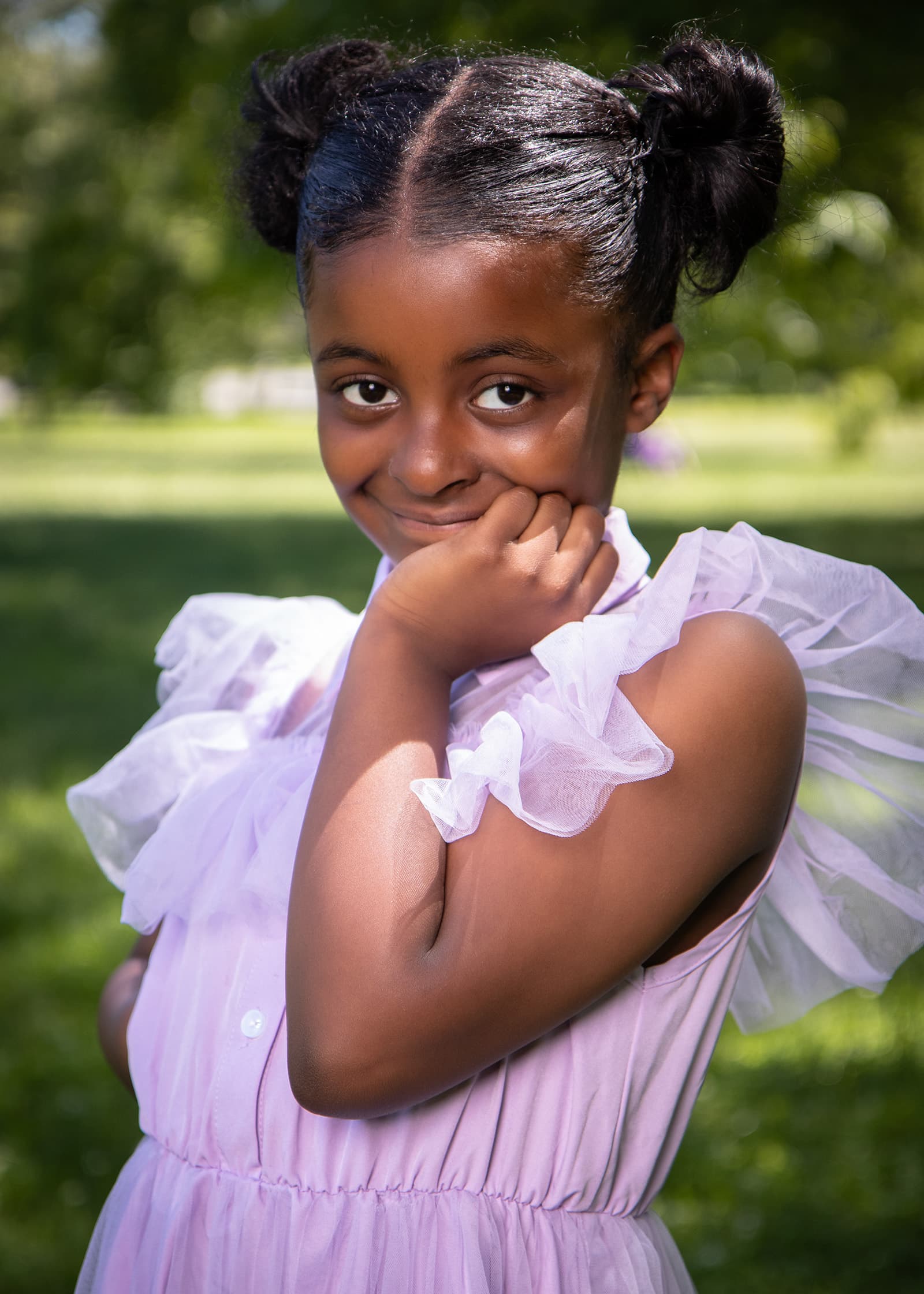 Outdoor portrait of a young girl in a lavender ruffled dress posing with her hand on her cheek in bright natural light.