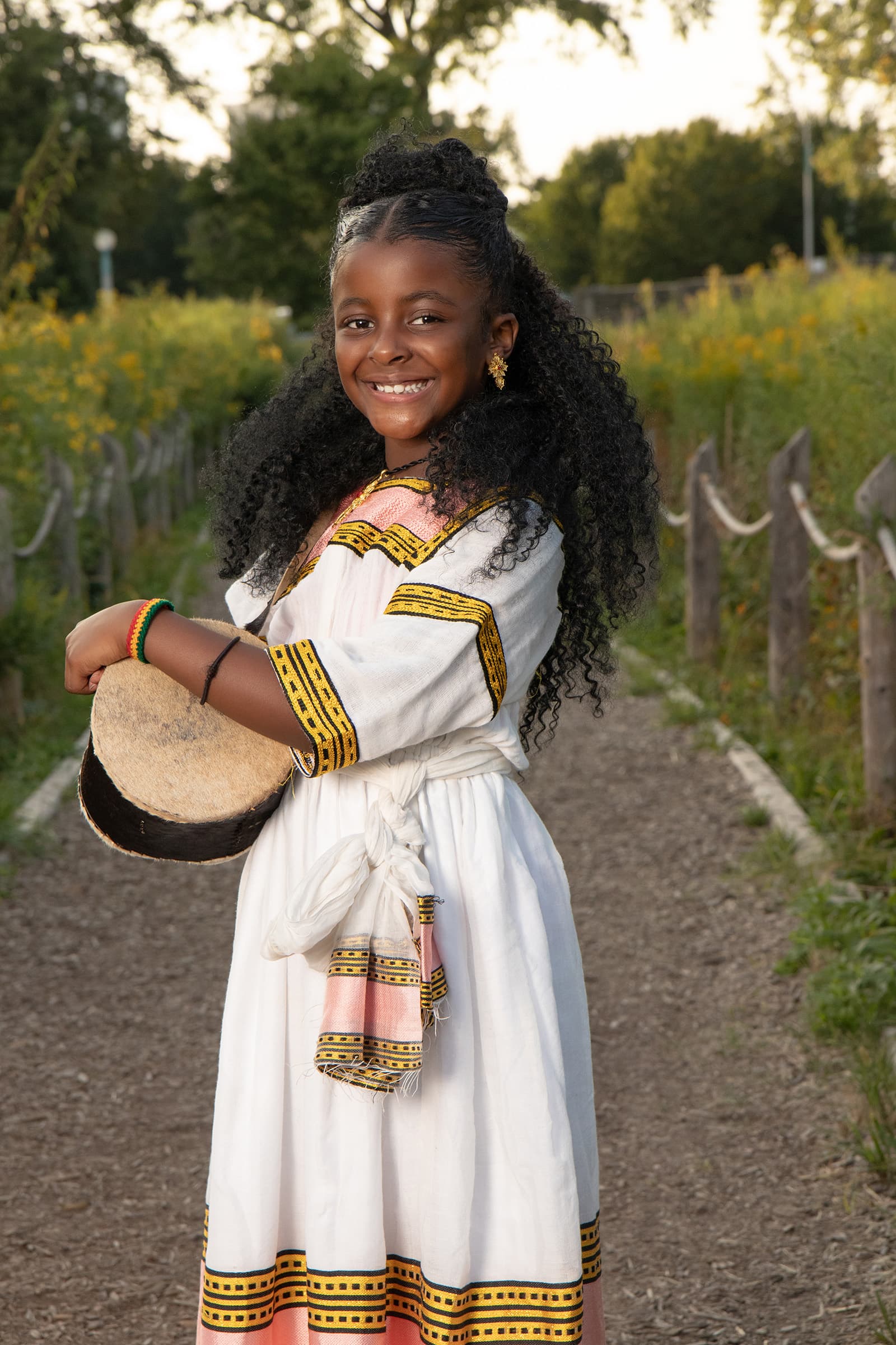 Outdoor portrait of a smiling young girl in a traditional embroidered dress holding a drum along a sunlit garden path.