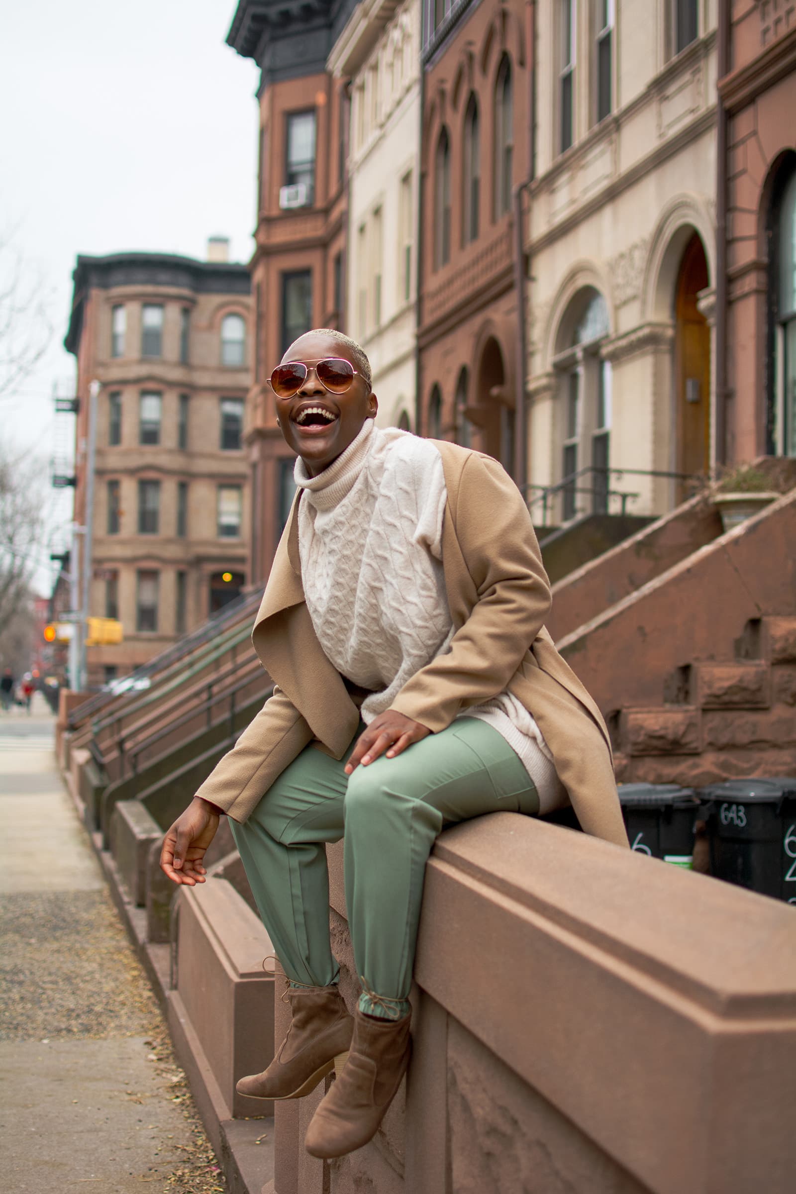 Lifestyle portrait of a smiling woman in a cream sweater and camel coat seated along a brownstone-lined urban street.