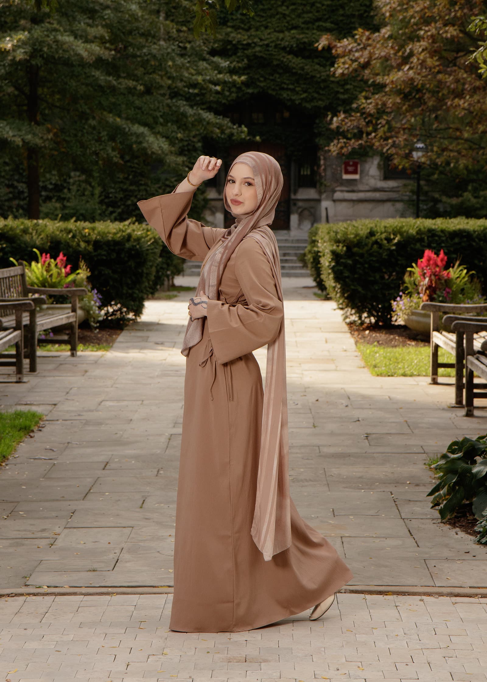 Full-length outdoor portrait of a woman in a taupe hijab and flowing neutral dress, standing along a formal garden walkway.