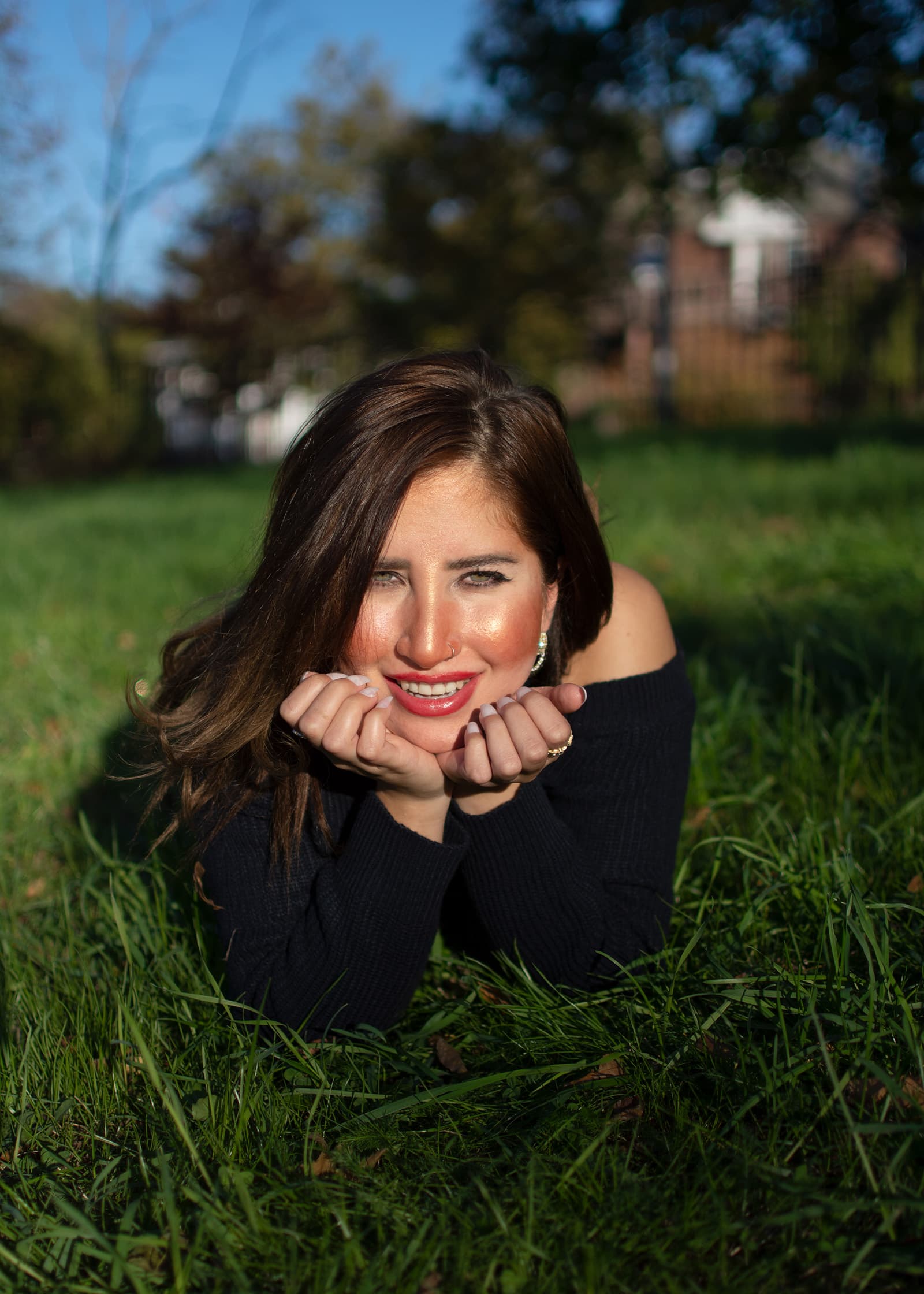 Outdoor portrait of a smiling woman lying in green grass, wearing a black off-shoulder top in warm natural sunlight.