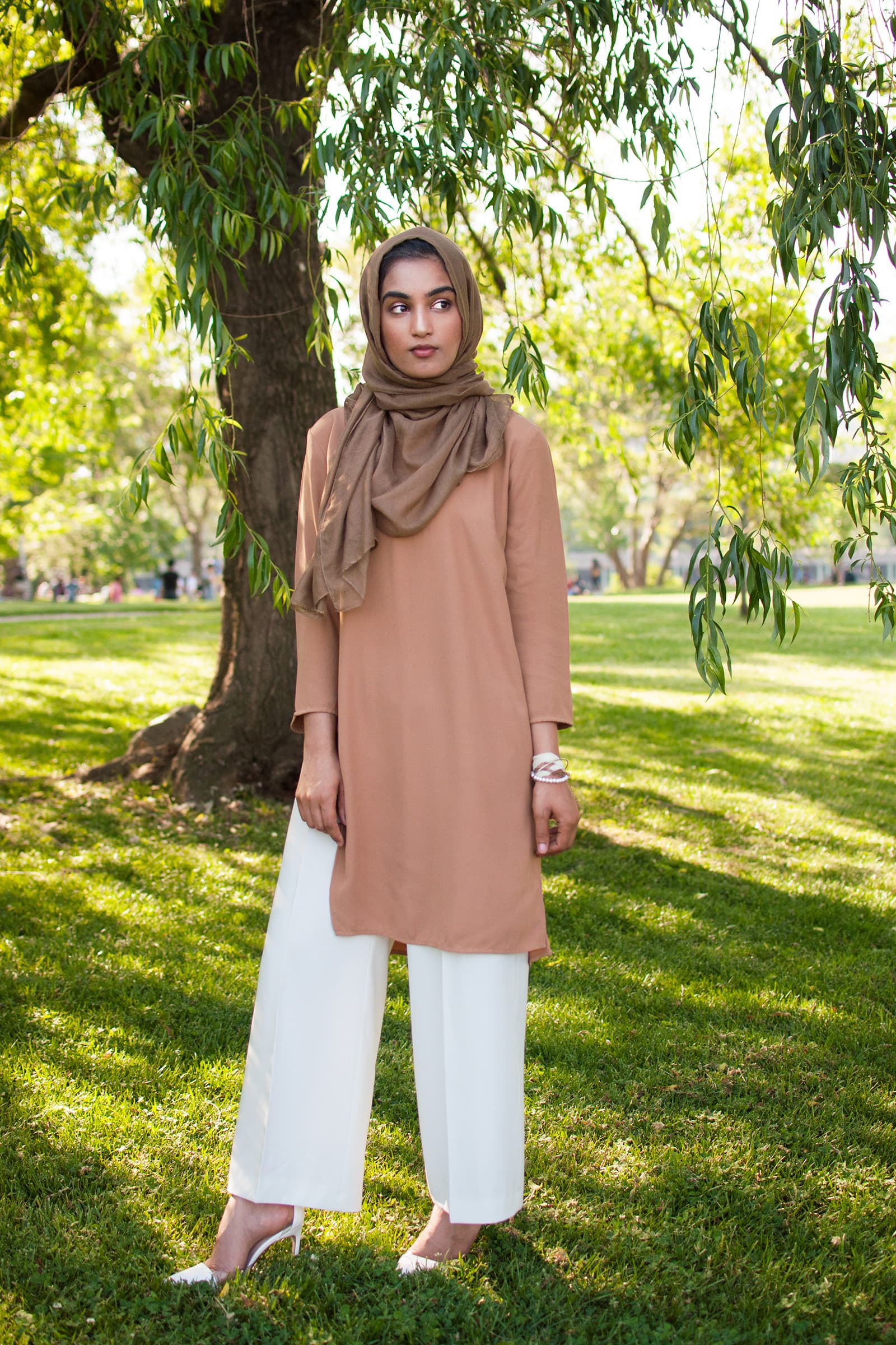 Full-length outdoor portrait of a woman in a brown hijab and neutral outfit standing beneath leafy trees in a park.