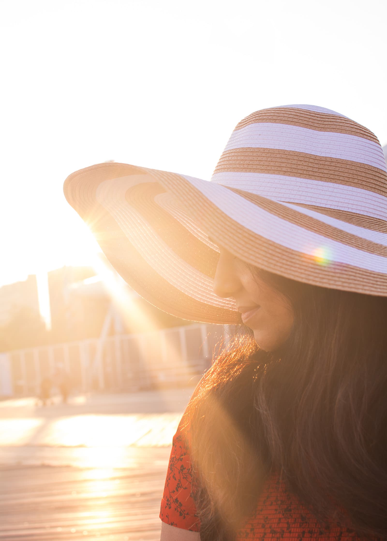 Backlit outdoor portrait of a woman wearing a wide-brim striped hat, photographed in warm golden-hour sunlight.
