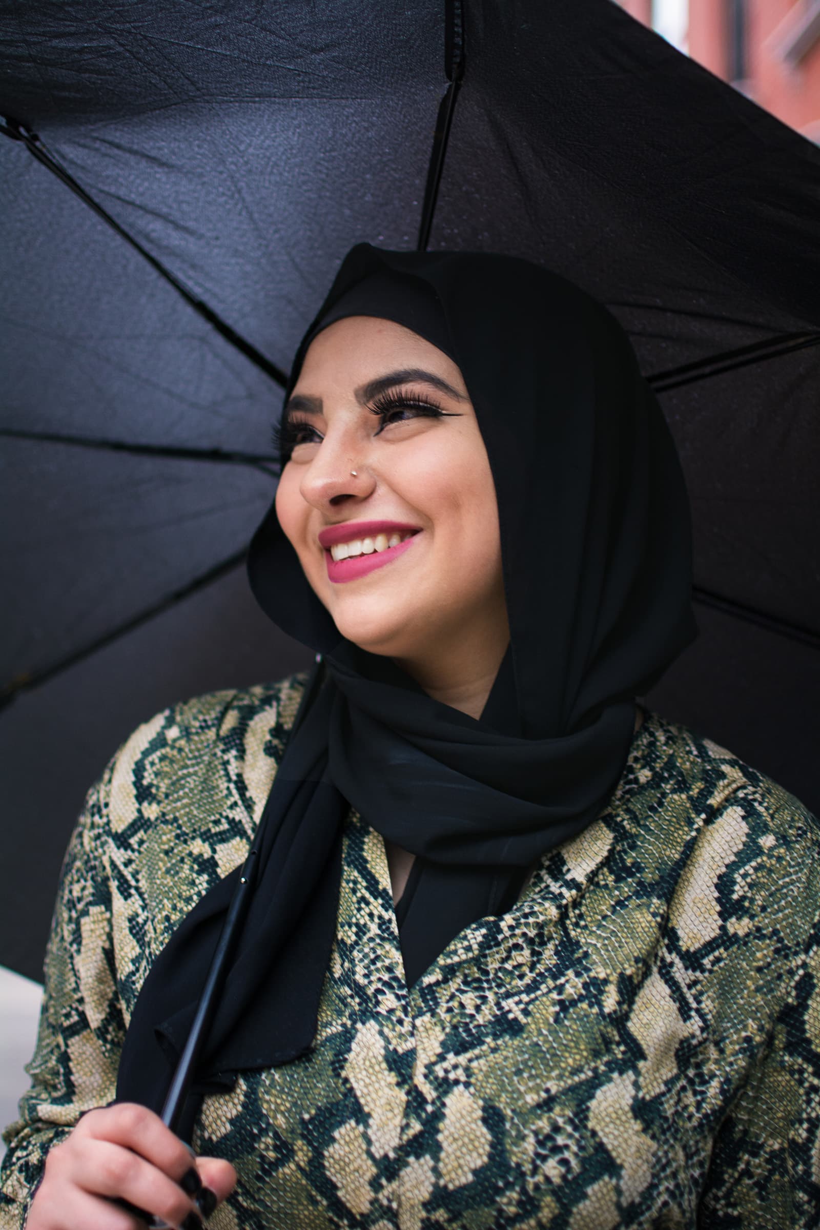 Close-up portrait of a smiling woman in a black hijab holding an umbrella, wearing a patterned jacket in soft daylight.