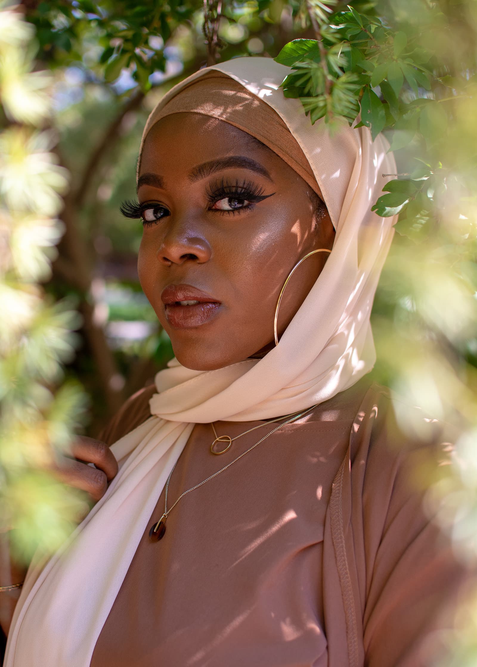 Close-up outdoor portrait of a woman in a cream hijab framed by green leaves and dappled sunlight in a natural setting.