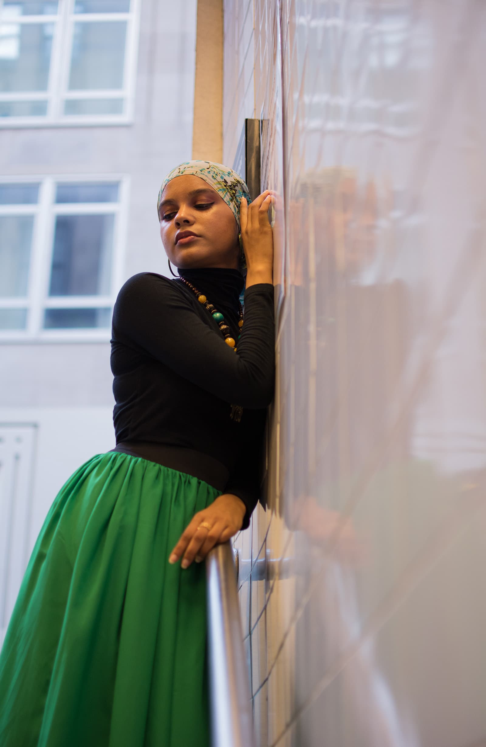 Indoor portrait of a woman in a floral headscarf, black top, and green skirt leaning against a reflective tiled wall.