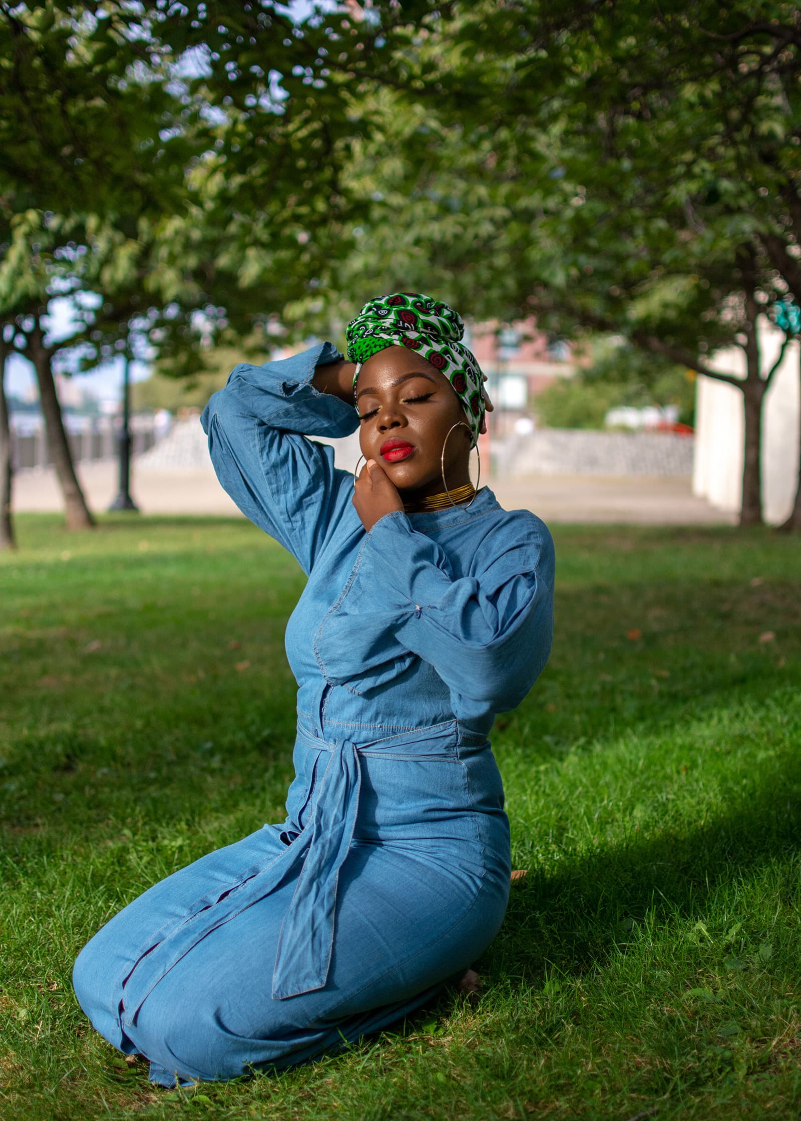 Outdoor portrait of a woman in a blue dress and patterned headwrap posing on grass beneath trees in soft sunlight.