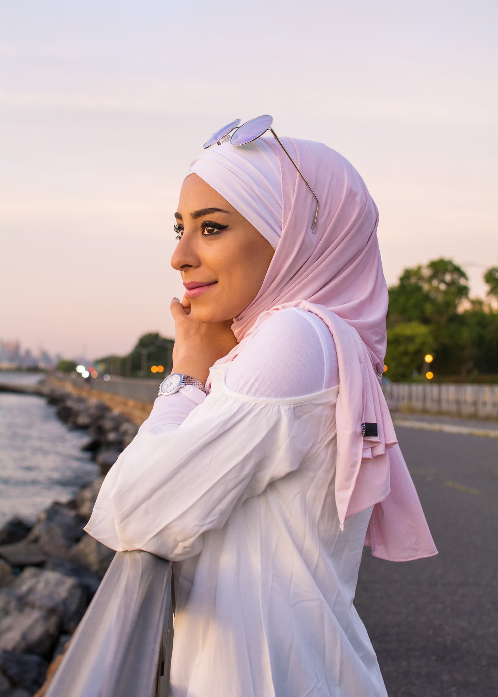 Golden-hour outdoor portrait of a woman in a pink hijab and white top posing beside a waterfront with soft evening light.