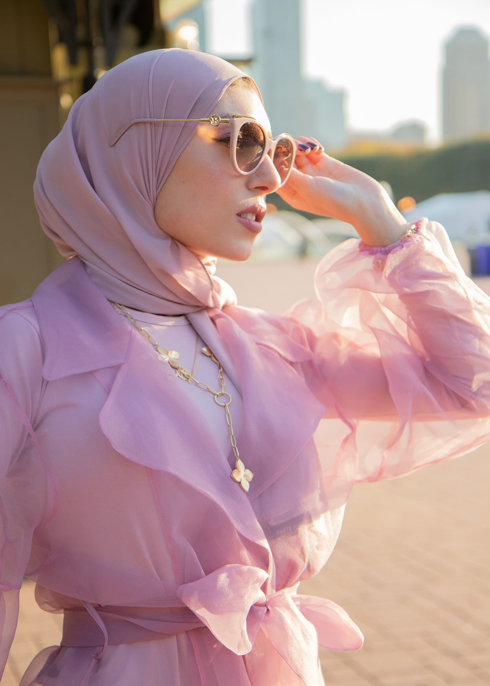 Golden-hour portrait of a woman in a pink hijab and sunglasses, wearing a flowing pink outfit with a city skyline behind her.