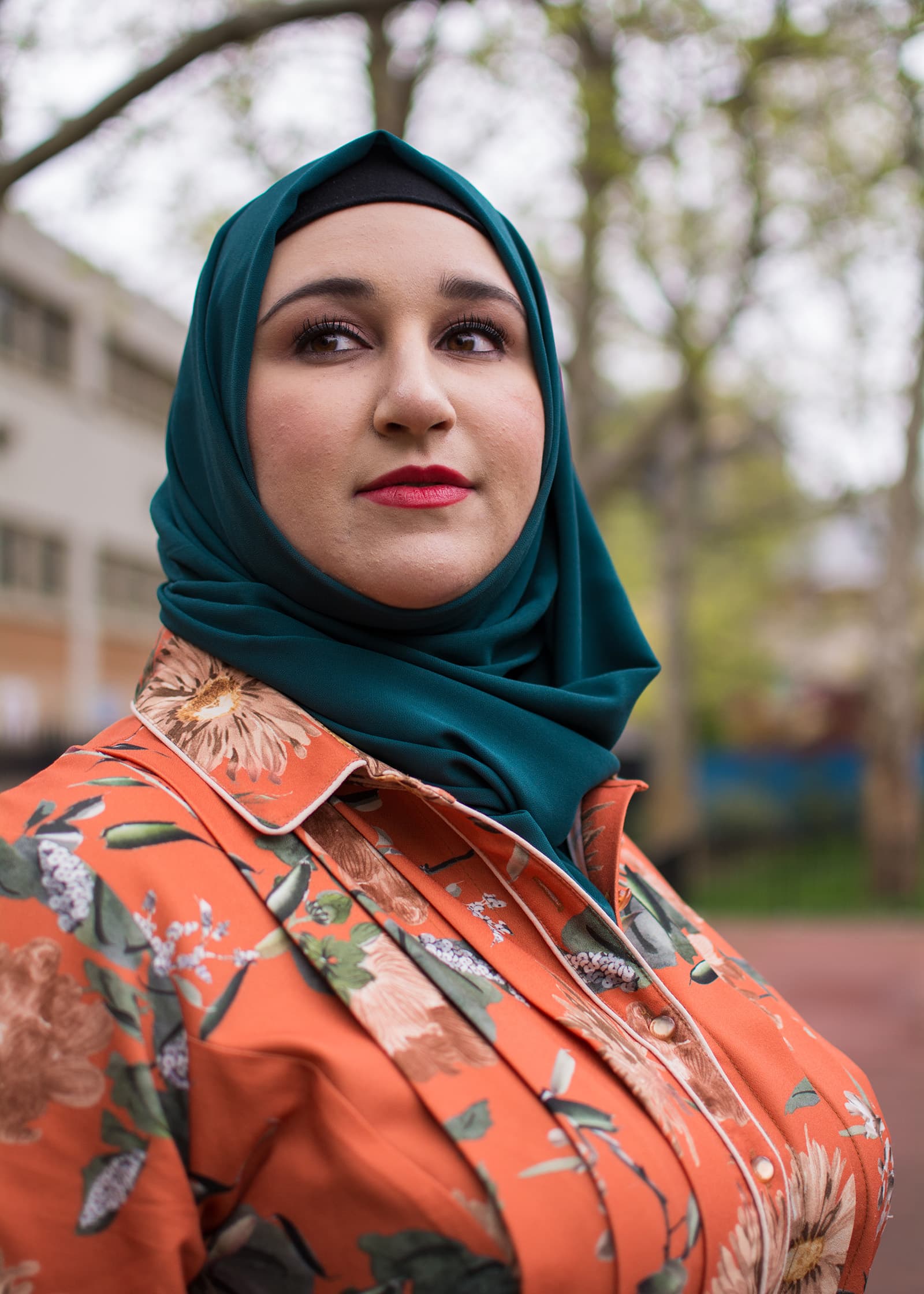 Outdoor portrait of a woman in a teal hijab and orange floral blouse, photographed with soft natural light and bokeh.