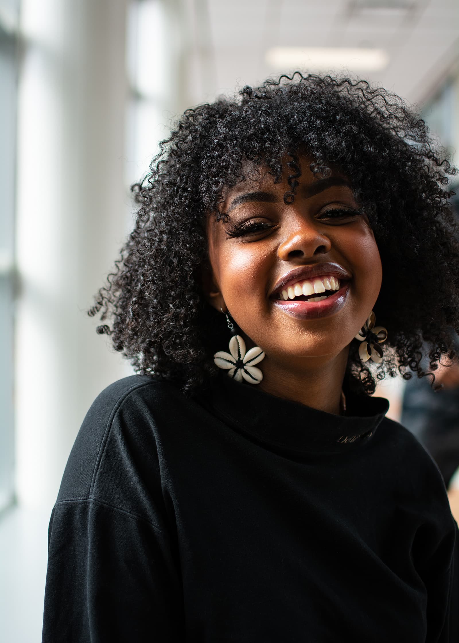 Indoor headshot of a smiling woman with natural curly hair and statement earrings, photographed in soft window light.