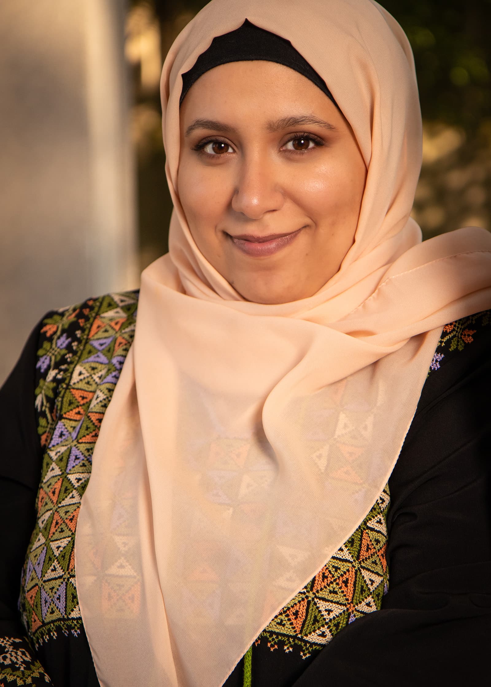 Close-up portrait of a woman in a peach hijab and embroidered black dress, photographed in warm natural light outdoors.