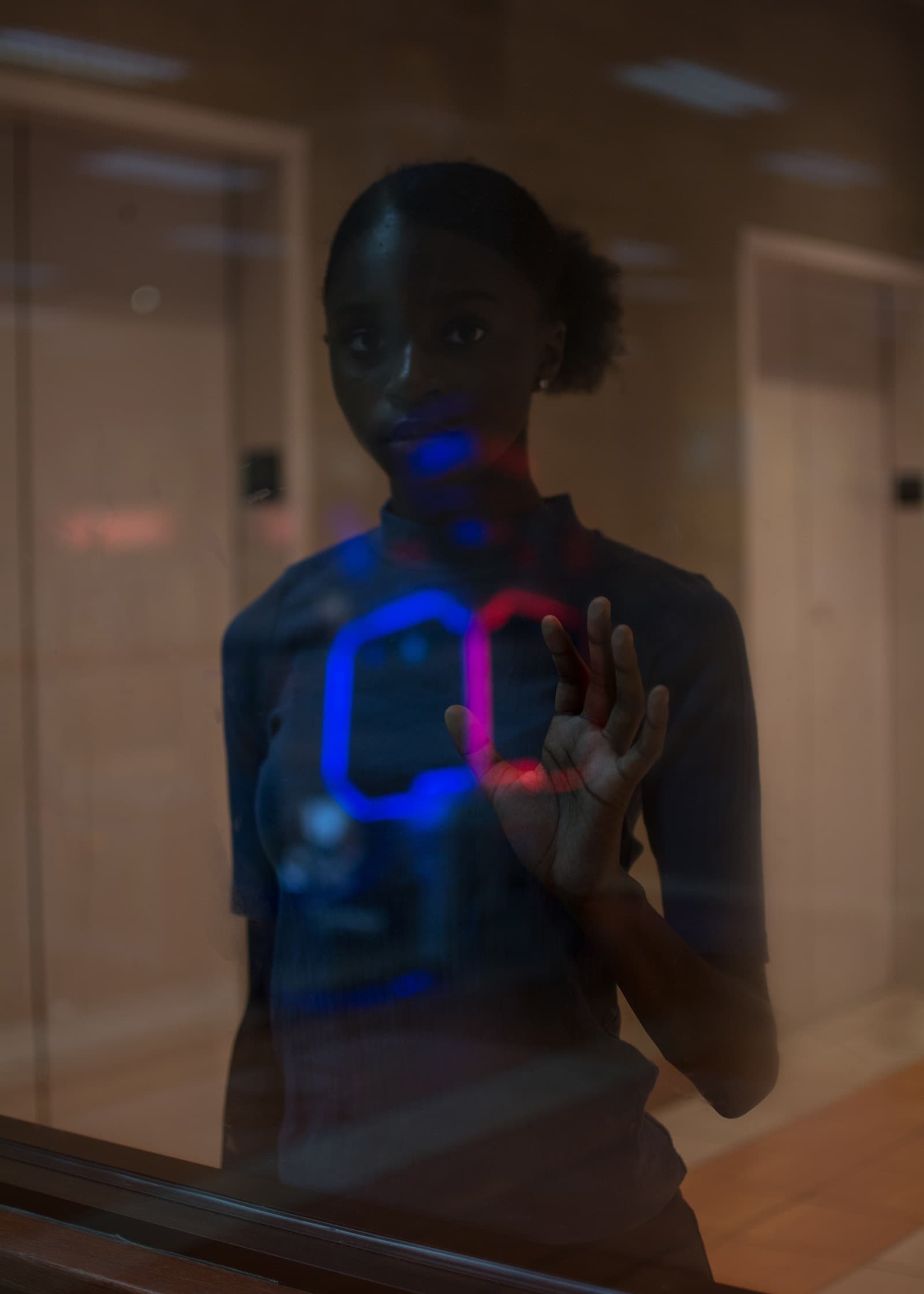 Moody indoor portrait of a woman behind reflective glass, with blue and red neon light creating an artistic layered effect.