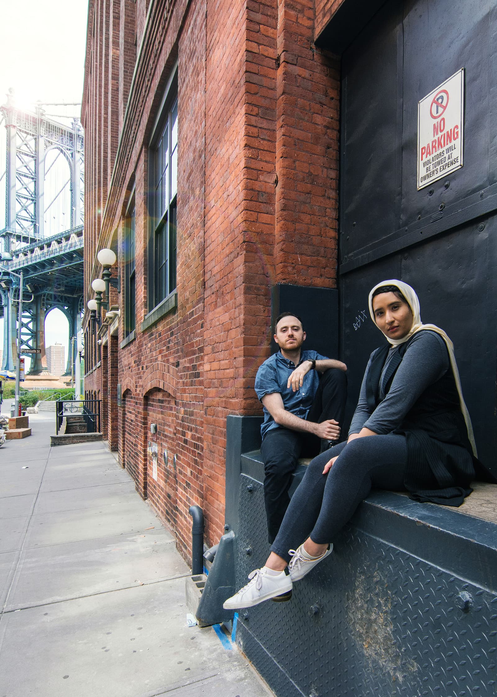 Environmental portrait of a man and woman seated beside a brick building near an urban bridge in natural daylight.