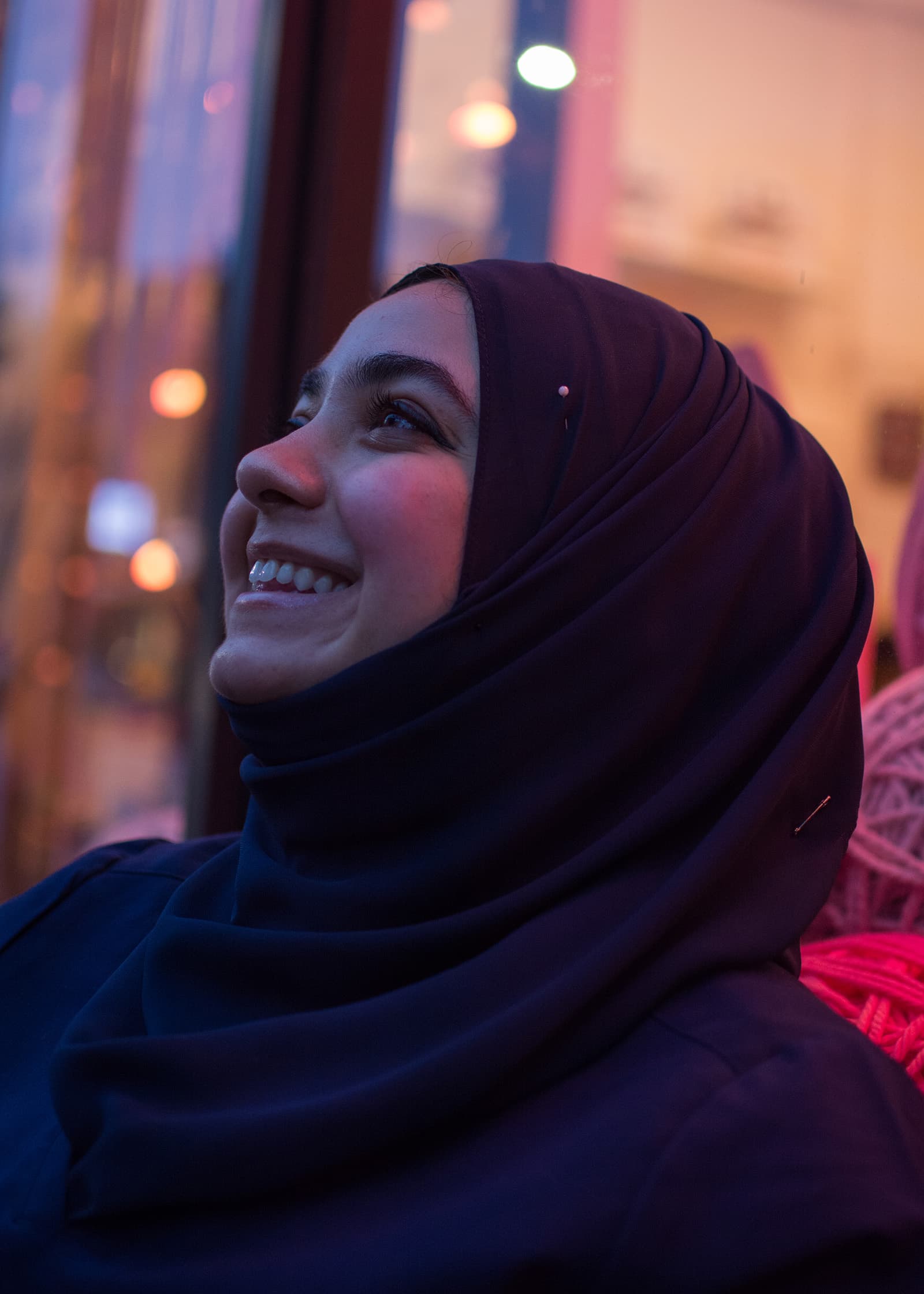 outdoor portrait of a smiling woman in a dark hijab, photographed beside a window with colorful ambient evening light.