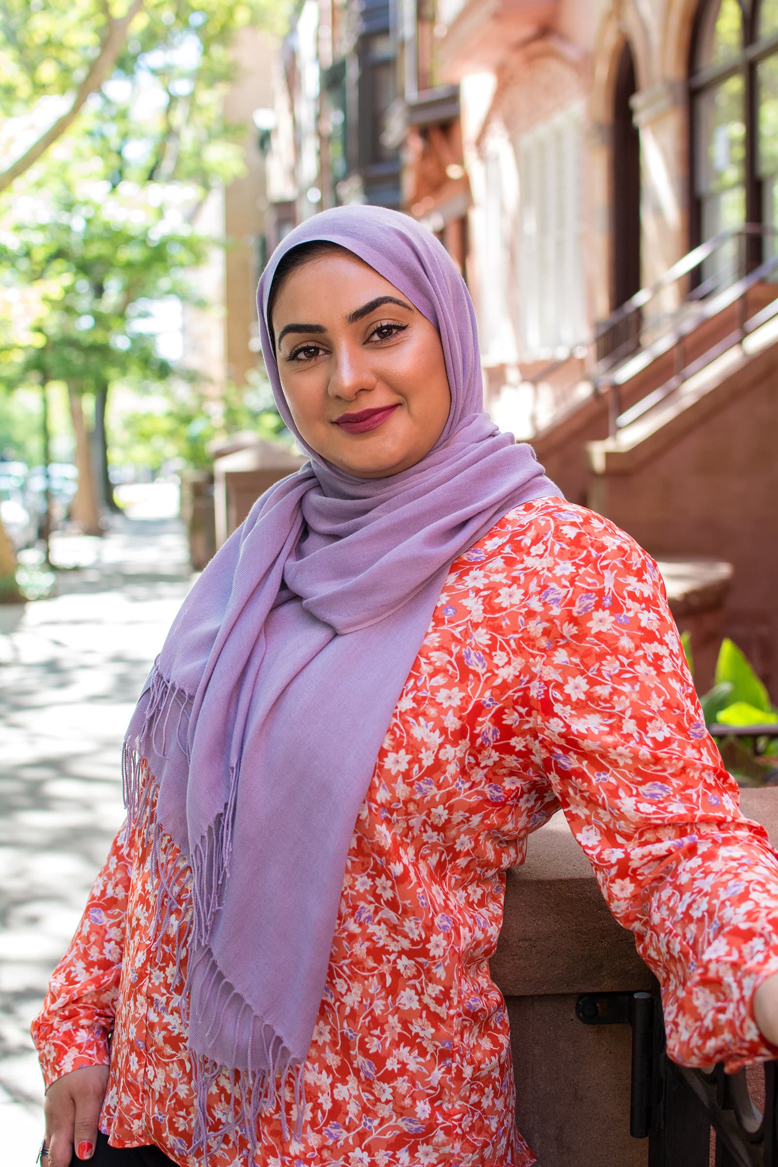 Outdoor portrait of a smiling woman in a lavender hijab and floral blouse, photographed on a tree-lined urban street.