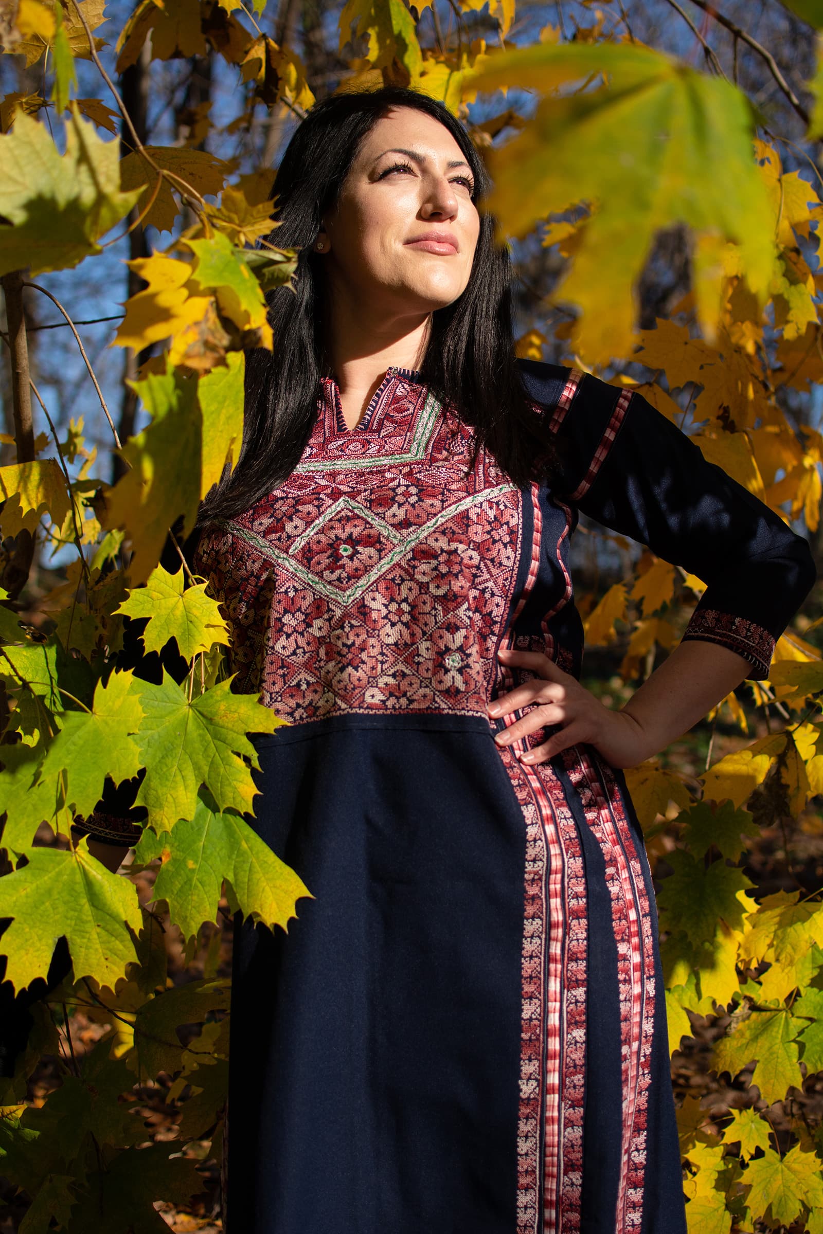 Outdoor portrait of a woman in a traditional patterned dress standing among autumn leaves in warm directional sunlight.