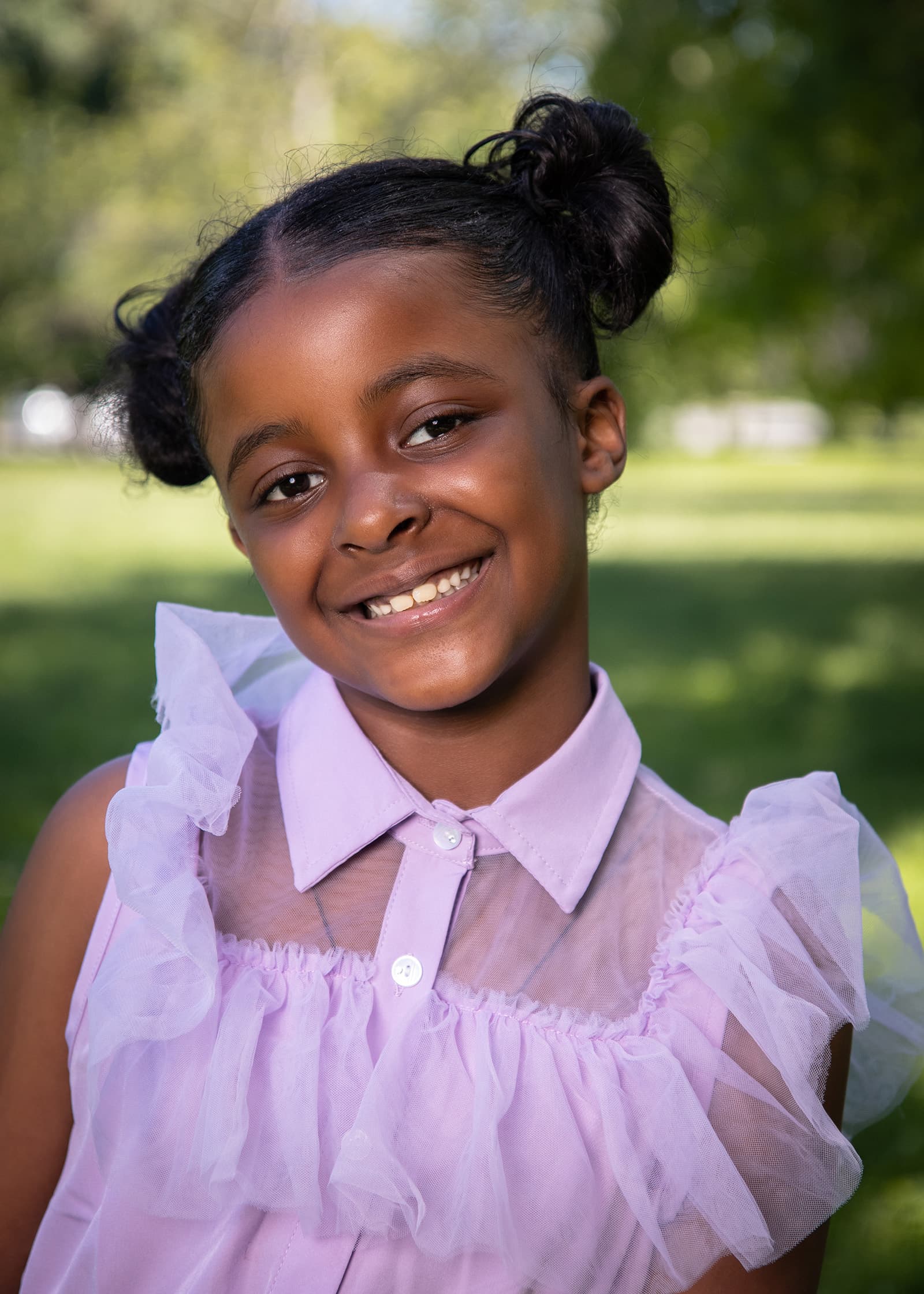 Outdoor portrait of a smiling young girl in a lavender ruffled dress, photographed in soft daylight in a green park.