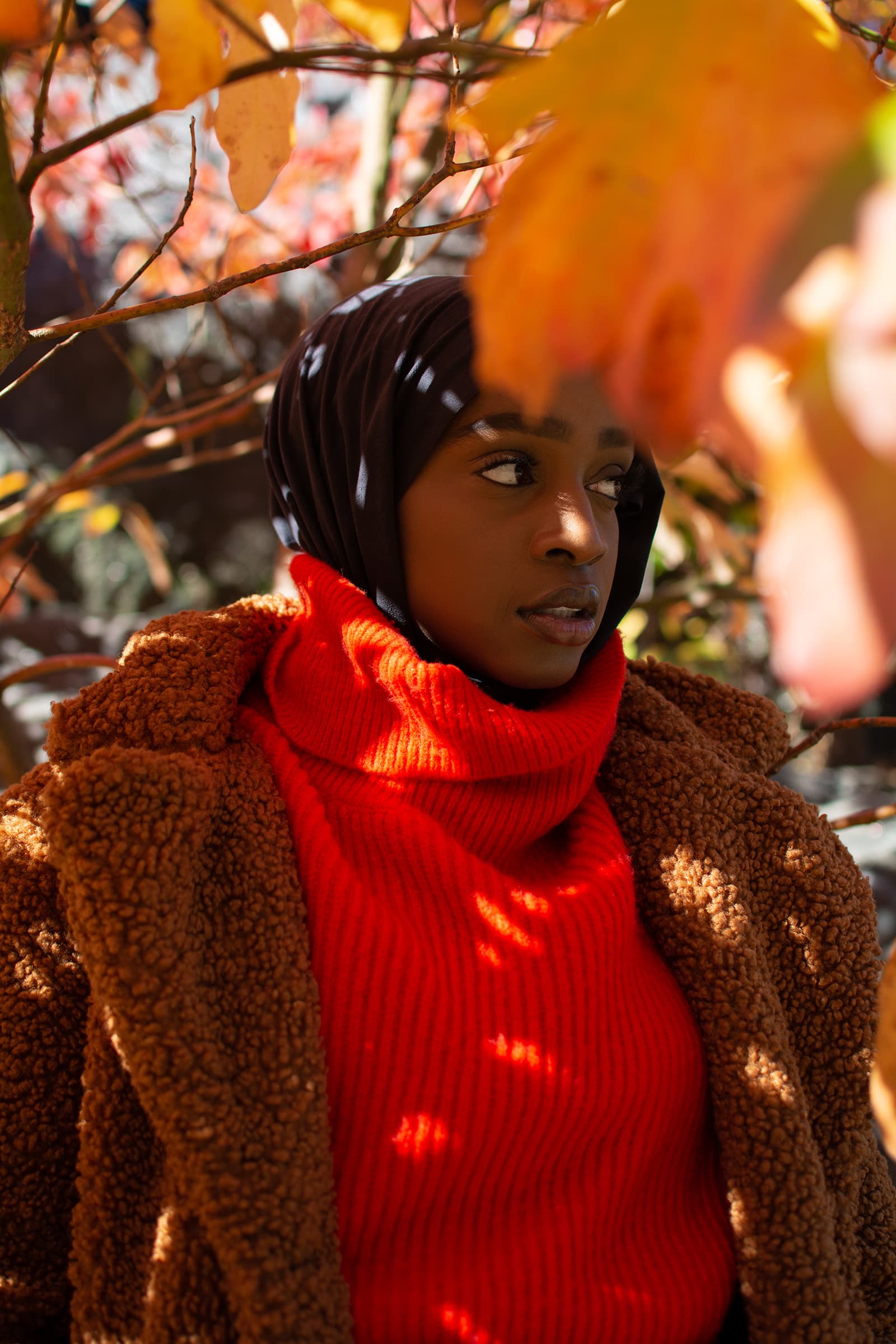 Outdoor portrait of a woman in a black headscarf and red sweater framed by autumn leaves in warm natural light.
