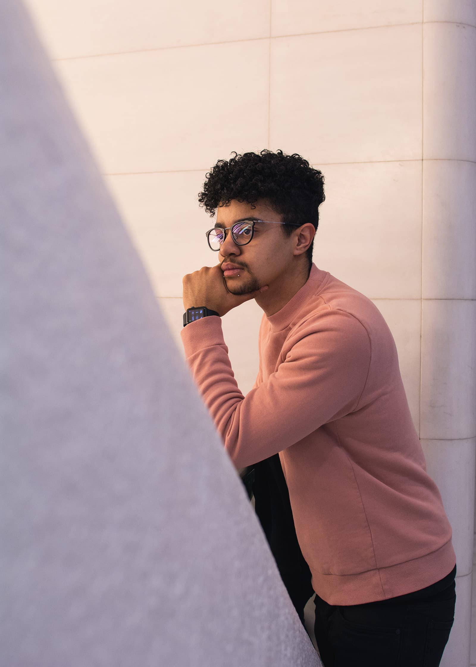 Environmental portrait of a man with curly hair and glasses wearing a salmon sweater beside modern architectural walls.