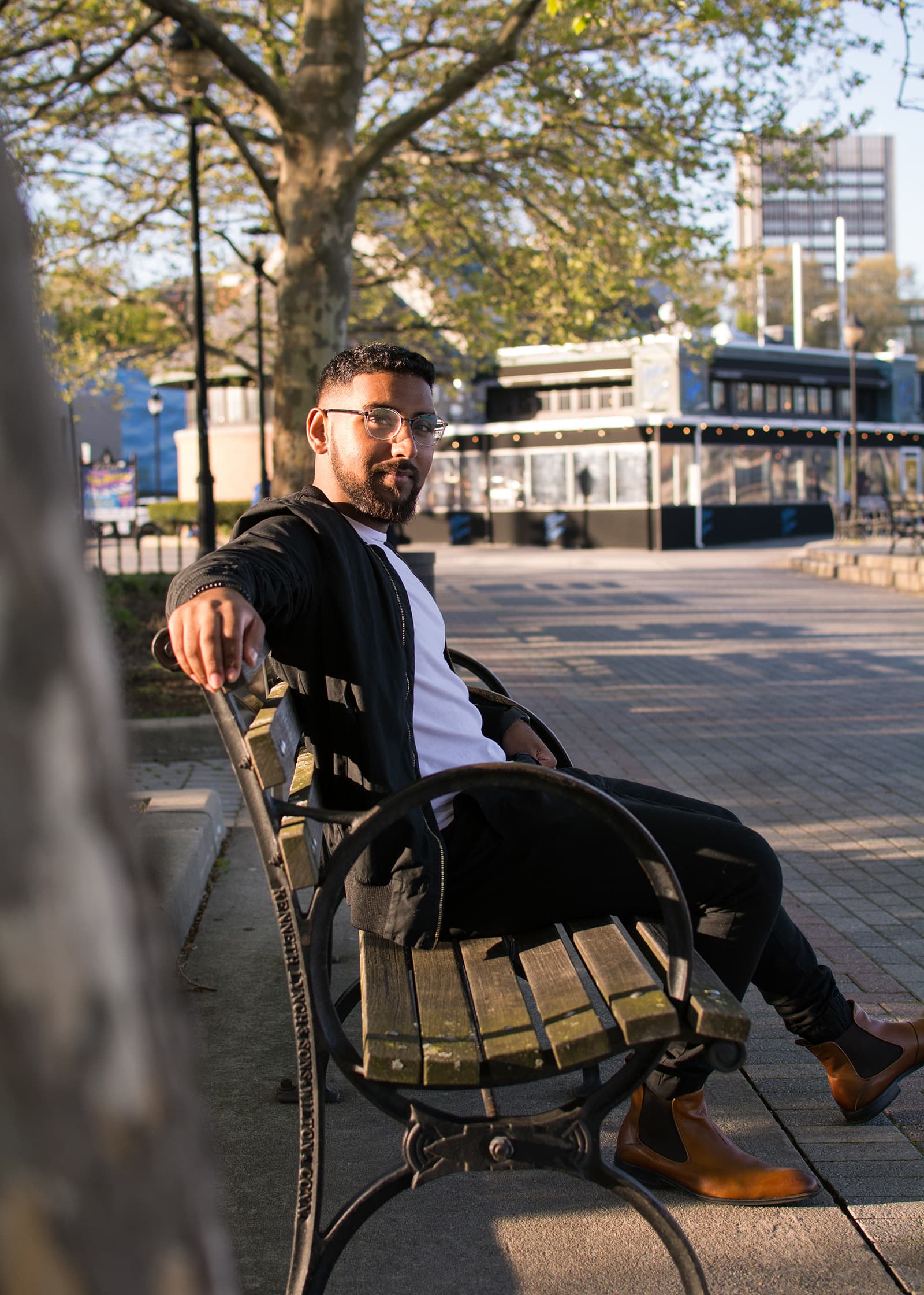 Outdoor portrait of a man with glasses and a beard seated on a park bench in warm late-afternoon natural light.