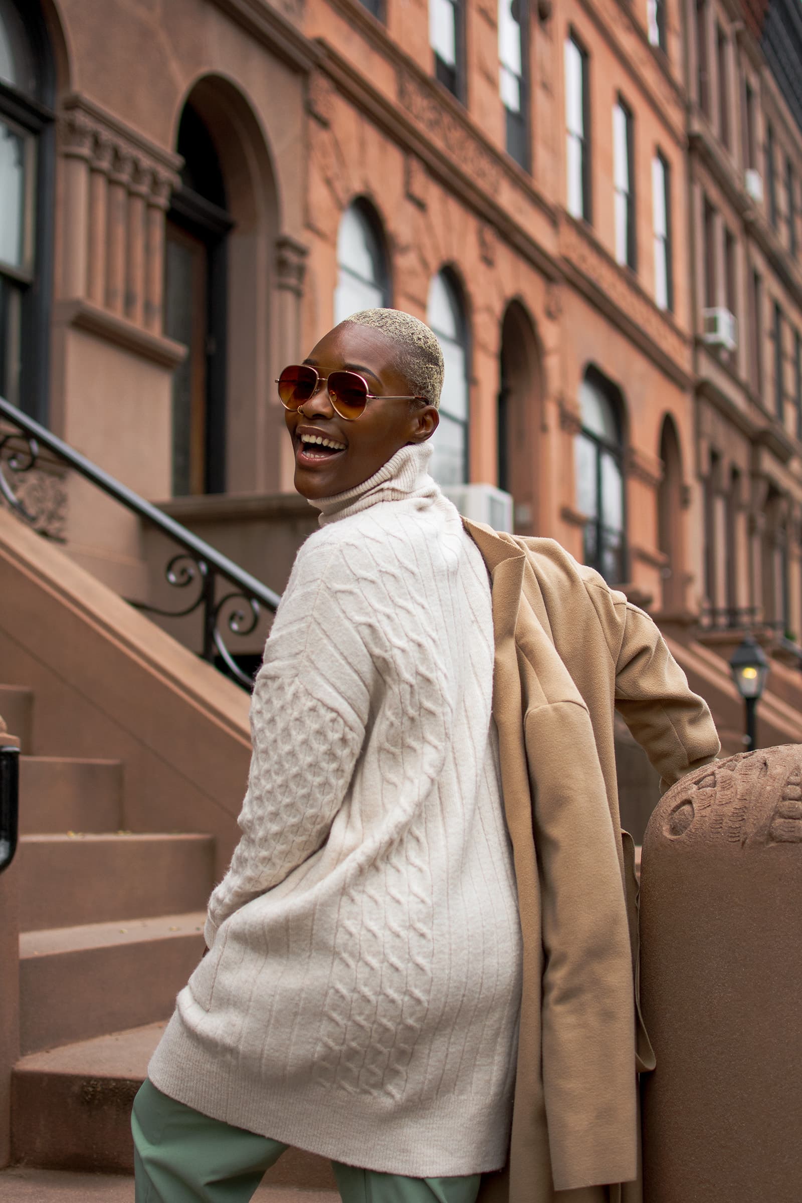 Outdoor portrait of a smiling woman with short blonde hair and sunglasses posing beside brownstone buildings in the city.