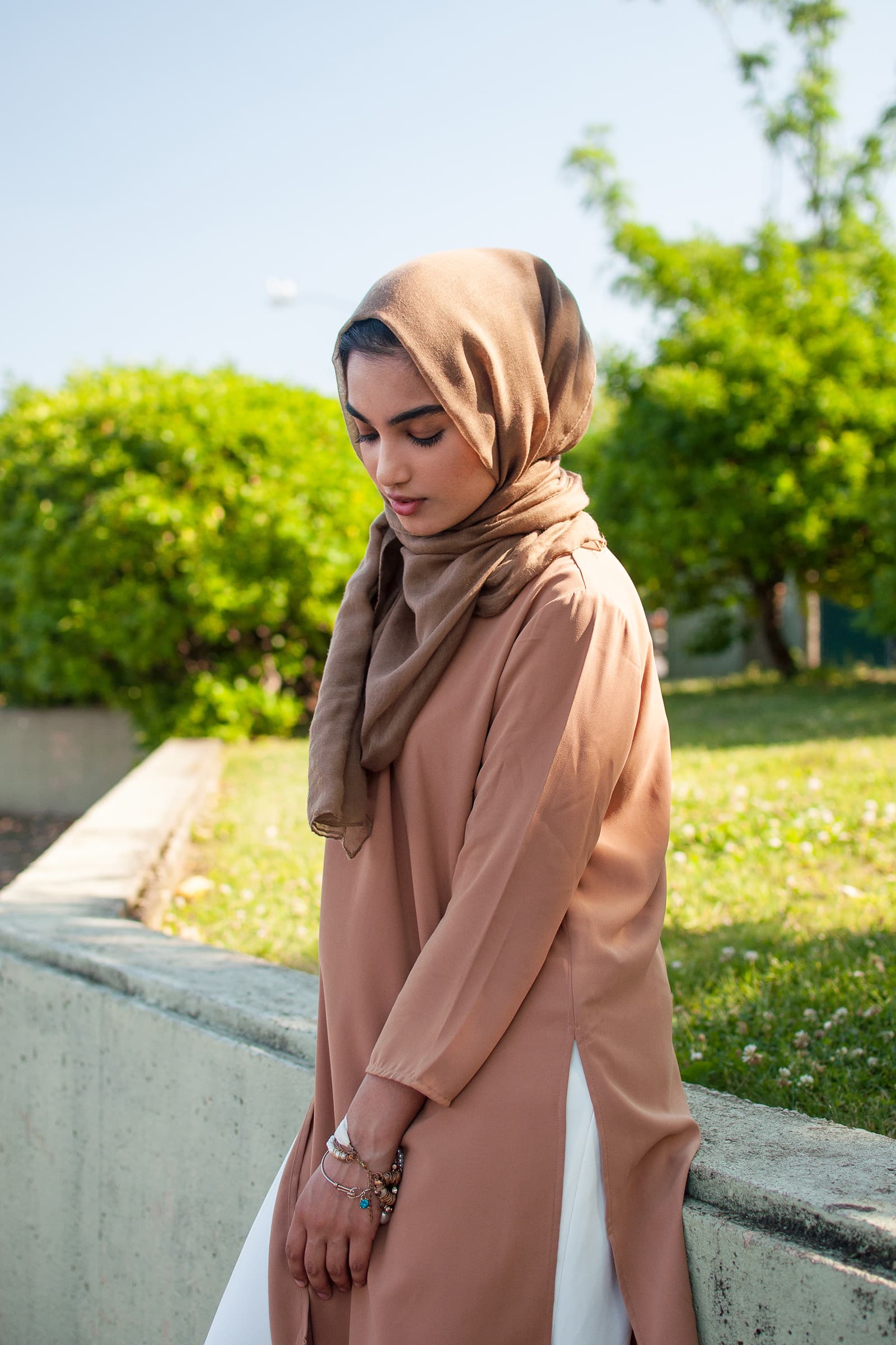 Outdoor portrait of a woman in a brown hijab and neutral outfit posing beside greenery in bright natural daylight.