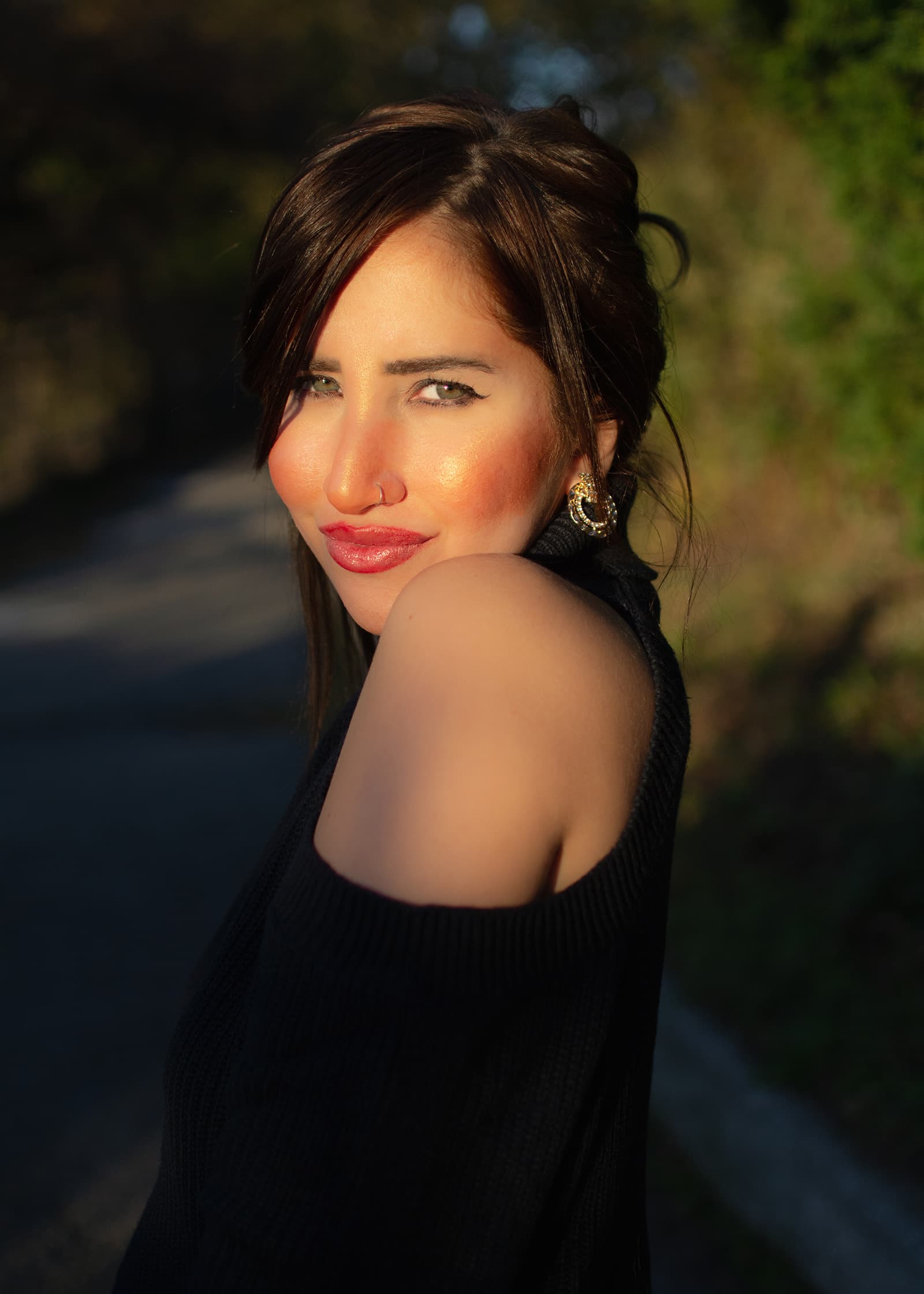 Outdoor portrait of a woman in a black off-shoulder top, photographed in warm golden-hour light against a blurred background.