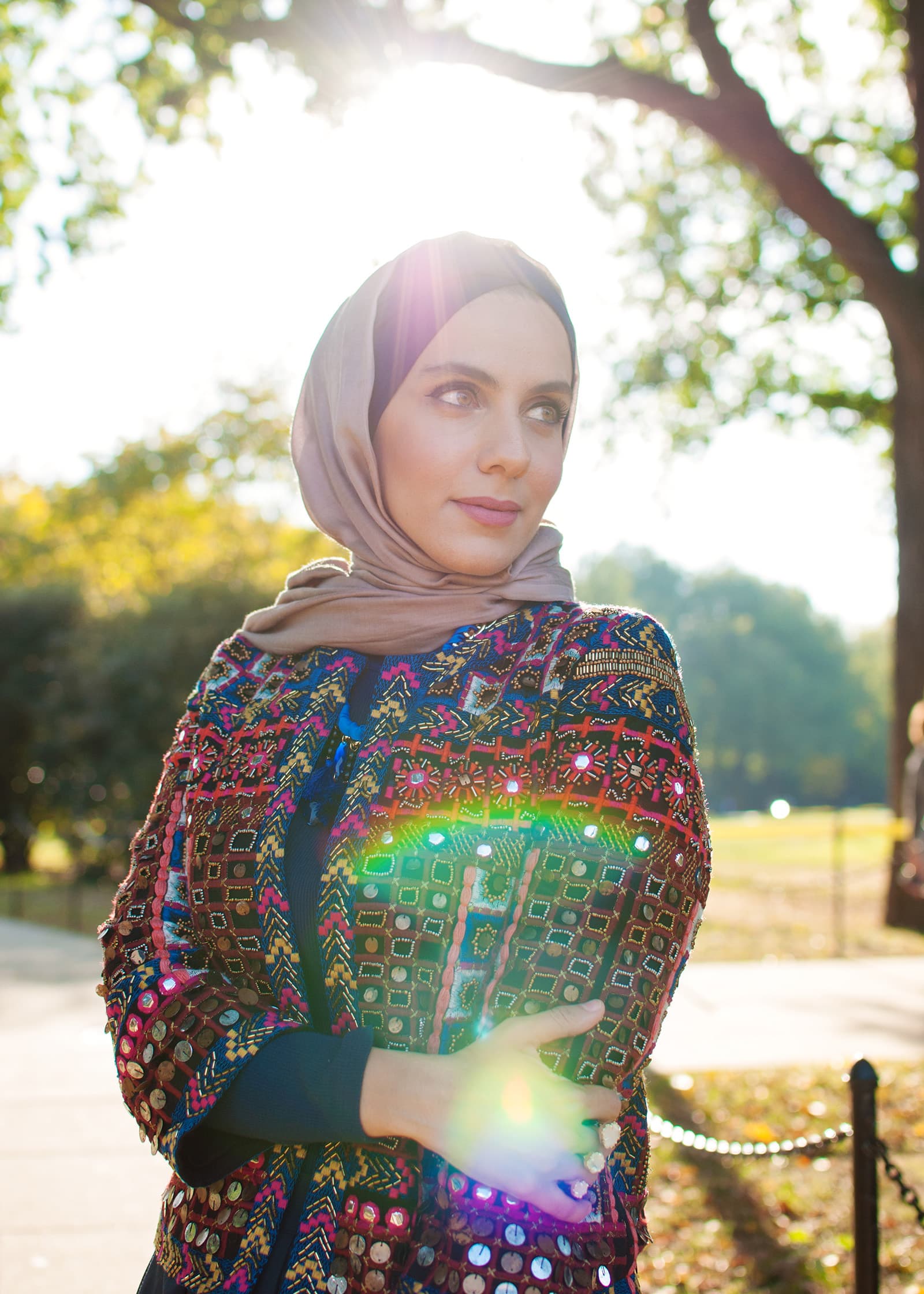 Outdoor portrait of a woman in a taupe hijab and colorful embroidered jacket, backlit by warm sunlight beneath leafy trees.