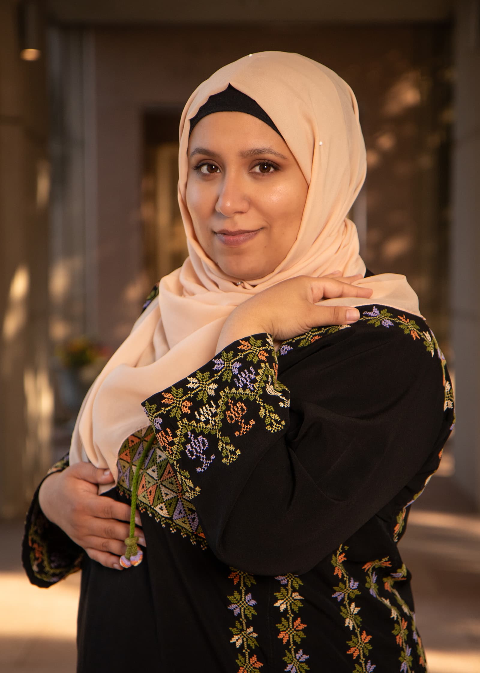 Outdoor portrait of a woman in a peach hijab and embroidered black dress, photographed in warm natural light with soft bokeh.