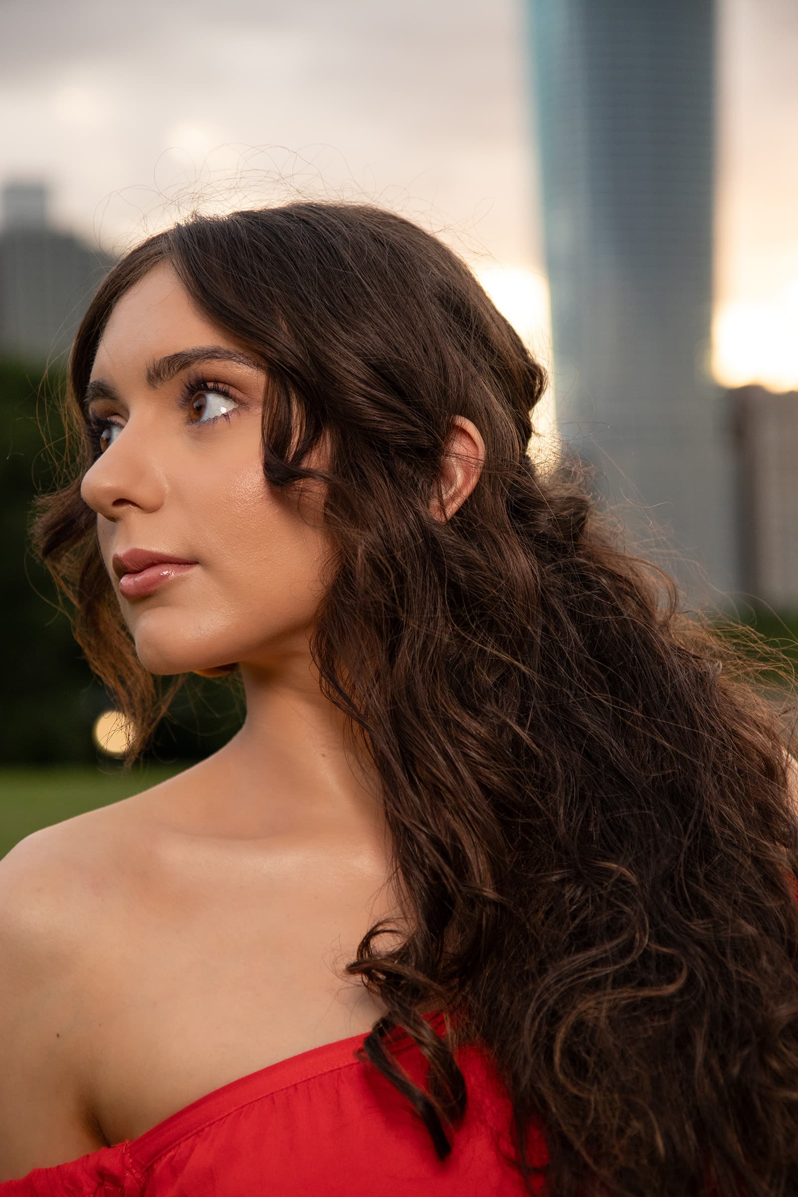 Outdoor beauty portrait of a woman in a red off-shoulder top with long dark curls, photographed at sunset with city skyline.