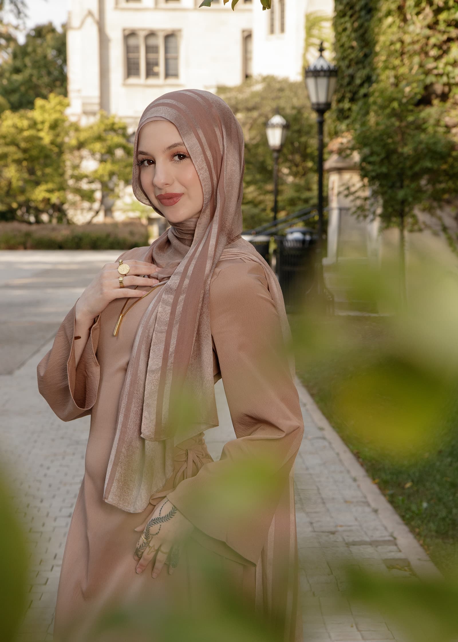 Outdoor portrait of a woman in a taupe hijab and matching dress, posed in a landscaped garden with soft natural light.