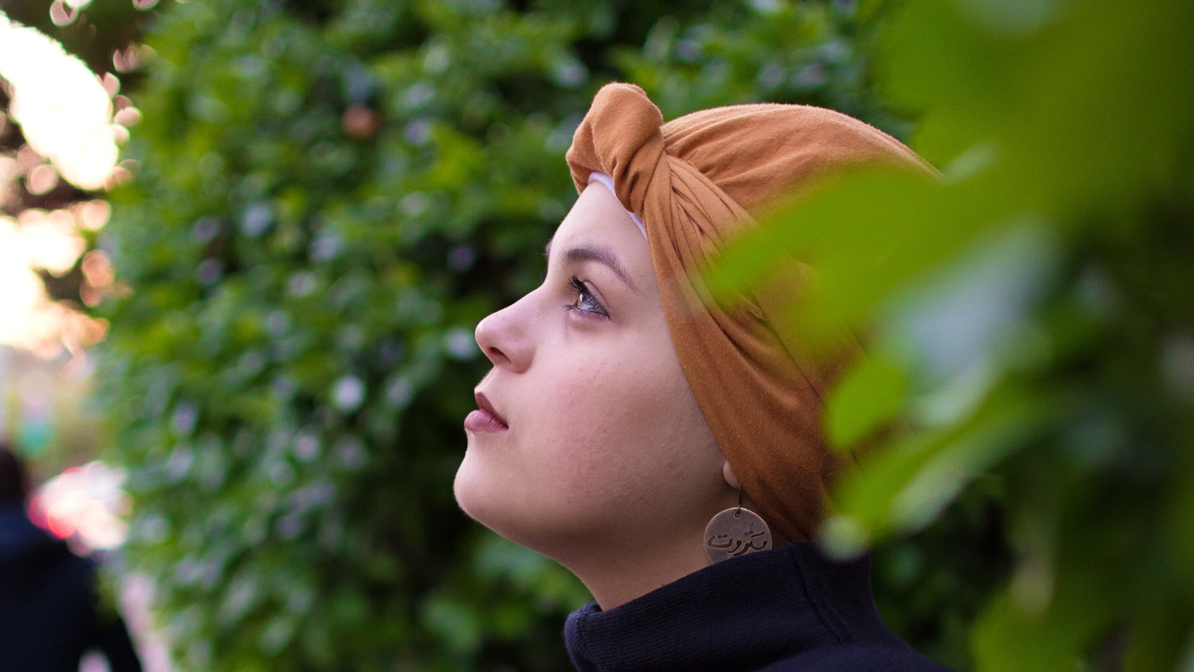 Outdoor cinematic portrait of a woman in a rust headwrap, framed by green foliage and photographed in soft natural light.
